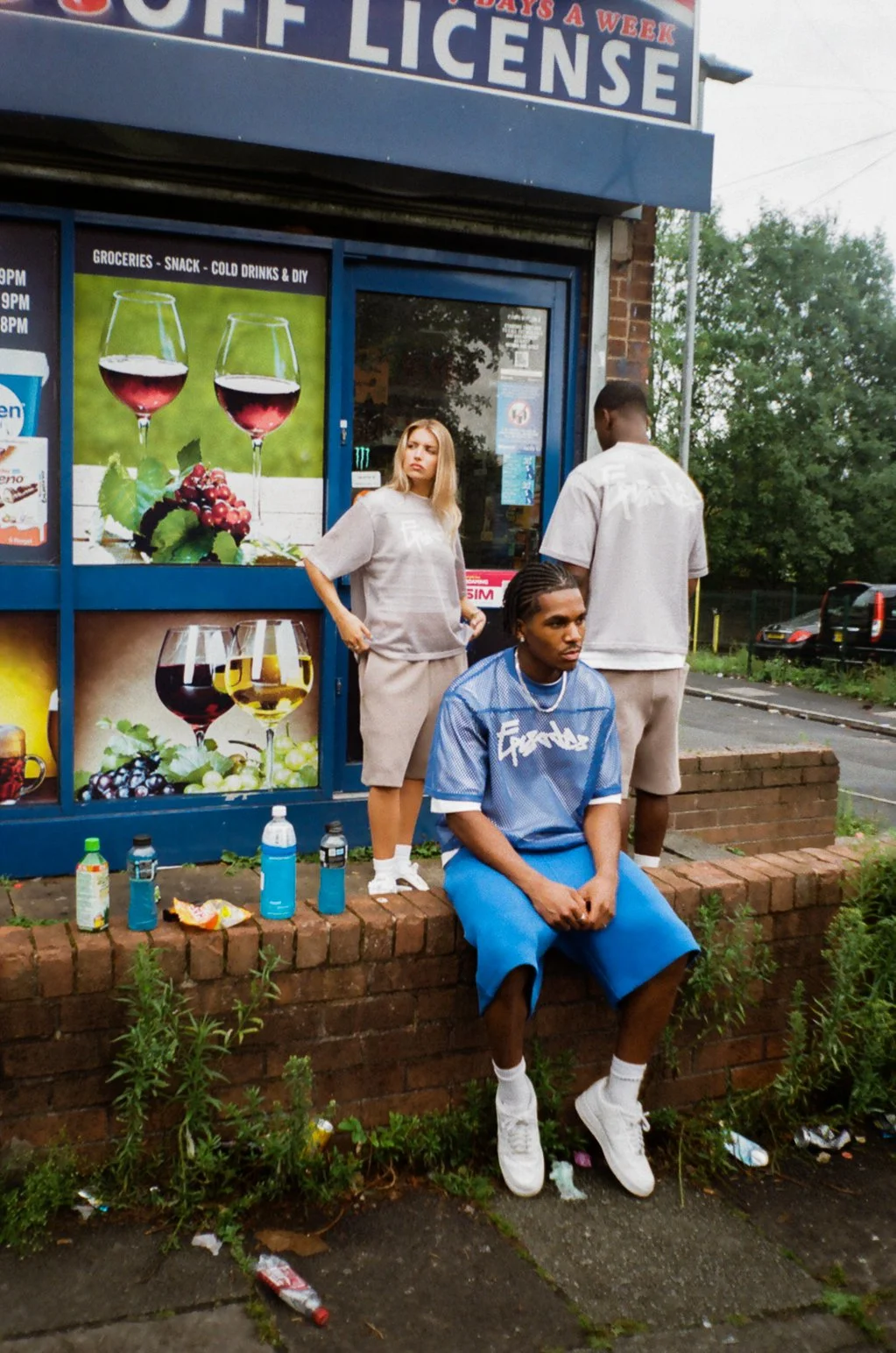 Three young people in front of a convenience store, with two men standing and one woman sitting on a brick ledge. The store has signage advertising alcohol, including wine and beer.