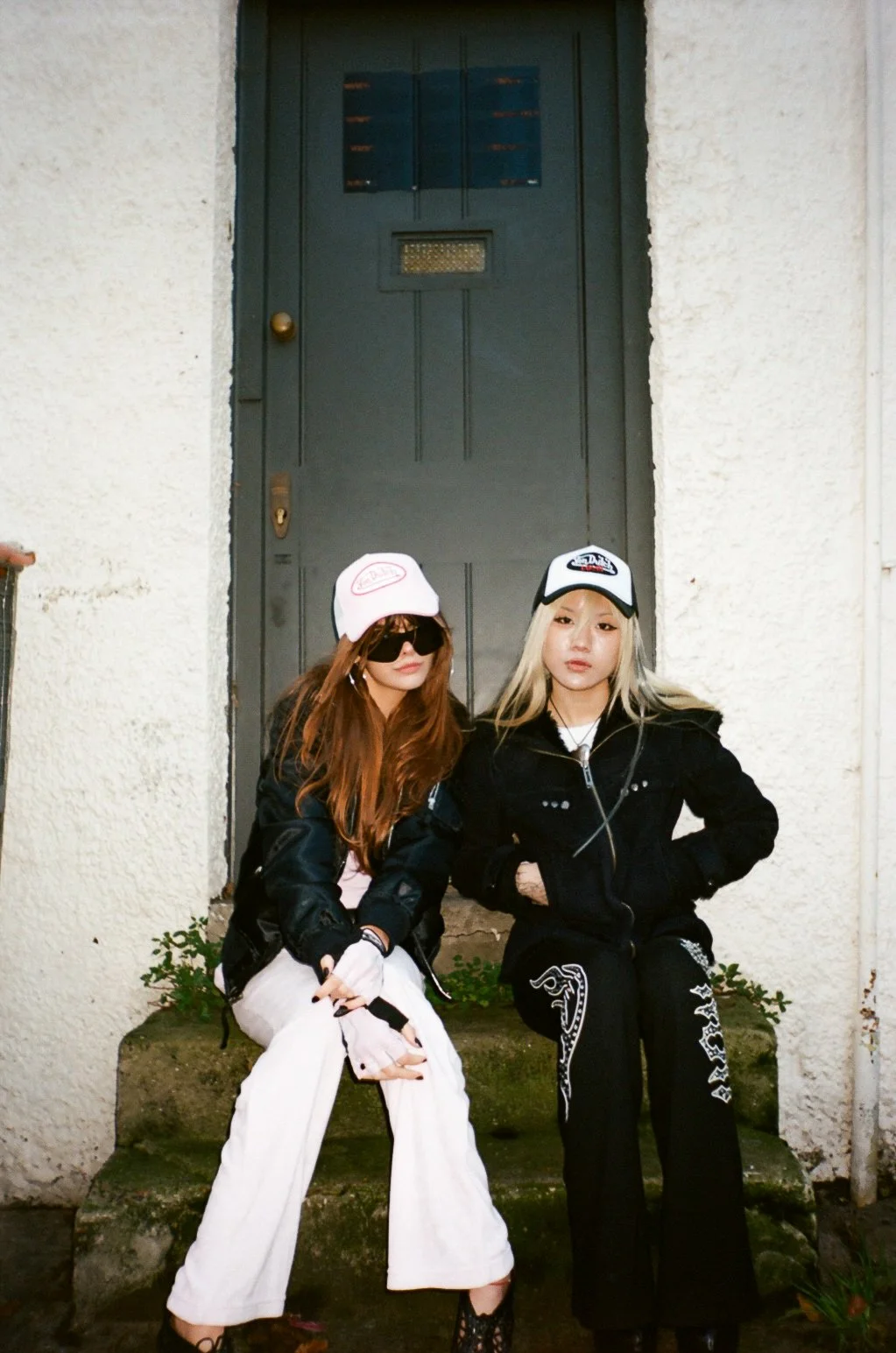Two young women sitting on steps in front of a black door with a mail slot. One woman has red hair, sunglasses, and is wearing a white cap, black jacket, and white pants. The other has blonde hair, a white cap, black jacket, and black pants with whit