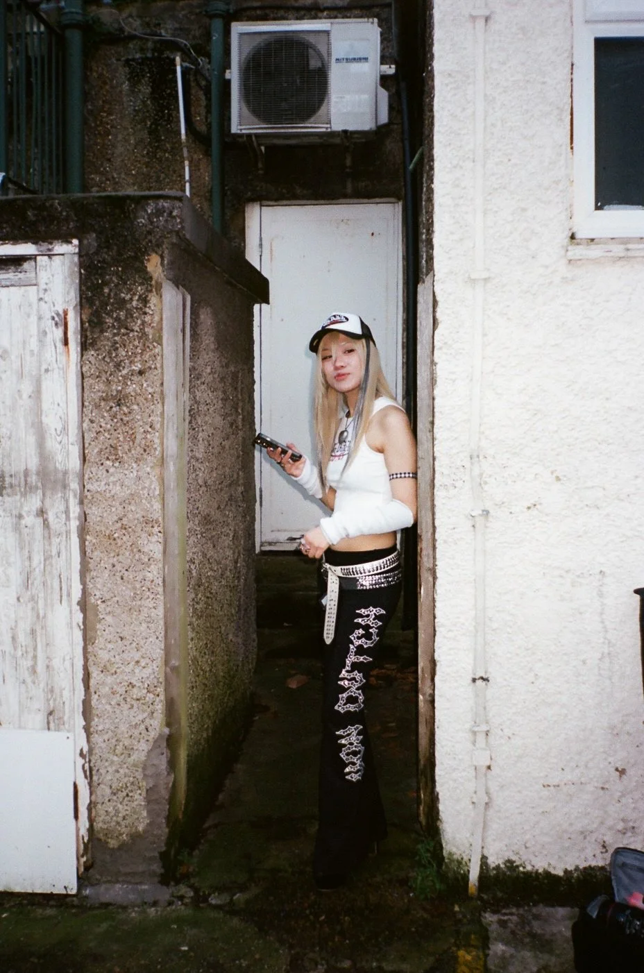A young woman with long blonde hair, wearing a white crop top, black pants with white designs, a baseball cap, and gloves, stands in a narrow alley holding a phone.