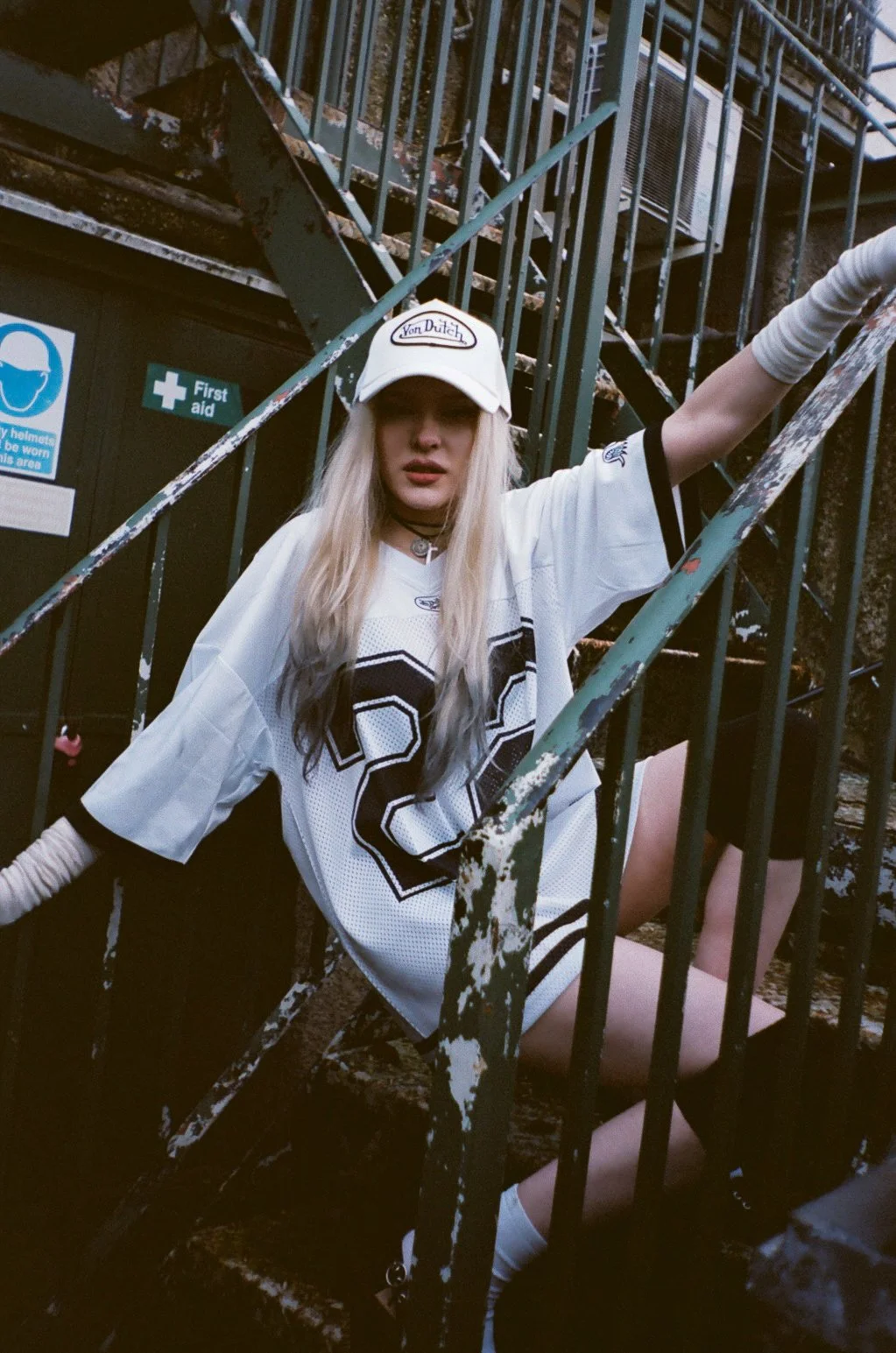 A young woman with long blonde hair sitting on a rusty outdoor metal staircase, wearing a white sports jersey with black accents, a white cap with 'Von Dutch' logo, and a choker necklace. She appears to be posing with her arm resting on the railing, 