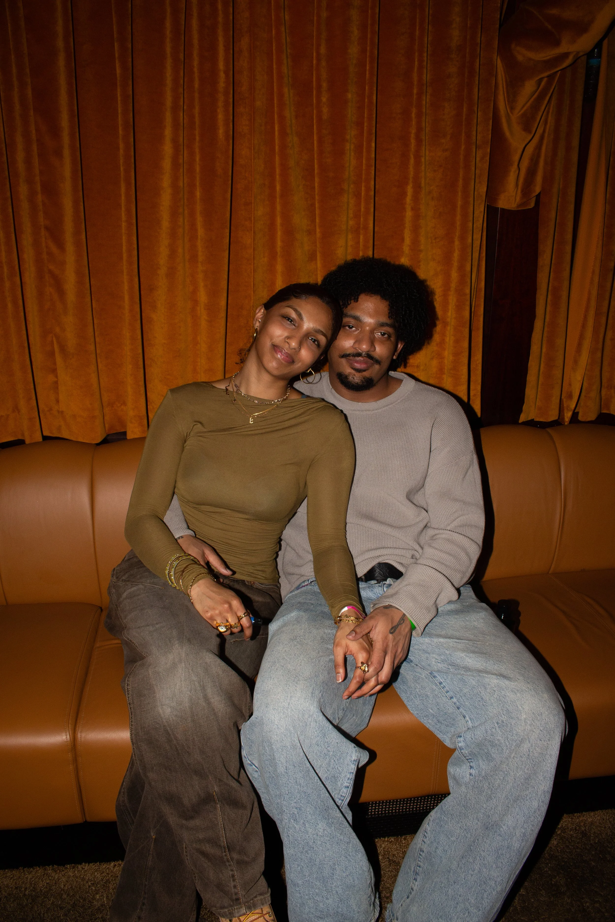 A woman and a man sit closely together on a tan sofa, smiling at the camera. The woman has her head tilted to the side, and the man has his arm around her. They are in front of brown curtains.