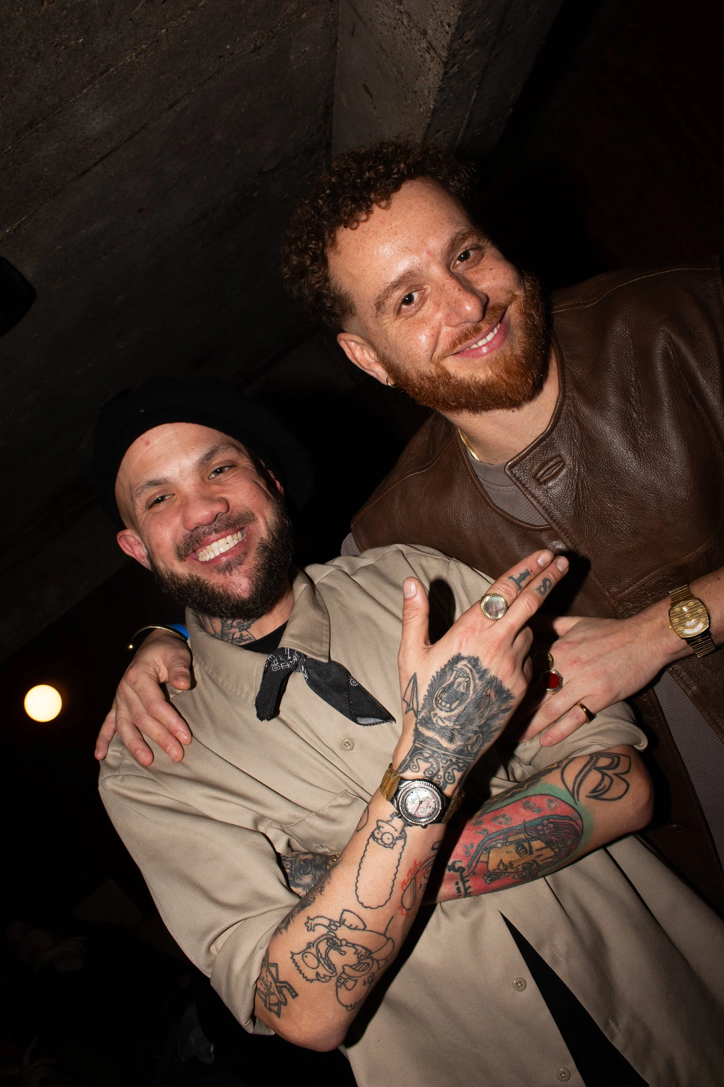 Two smiling men with tattoos, one wearing a beige shirt and black hat, the other in a brown leather jacket, posing closely together in a dimly lit setting.
