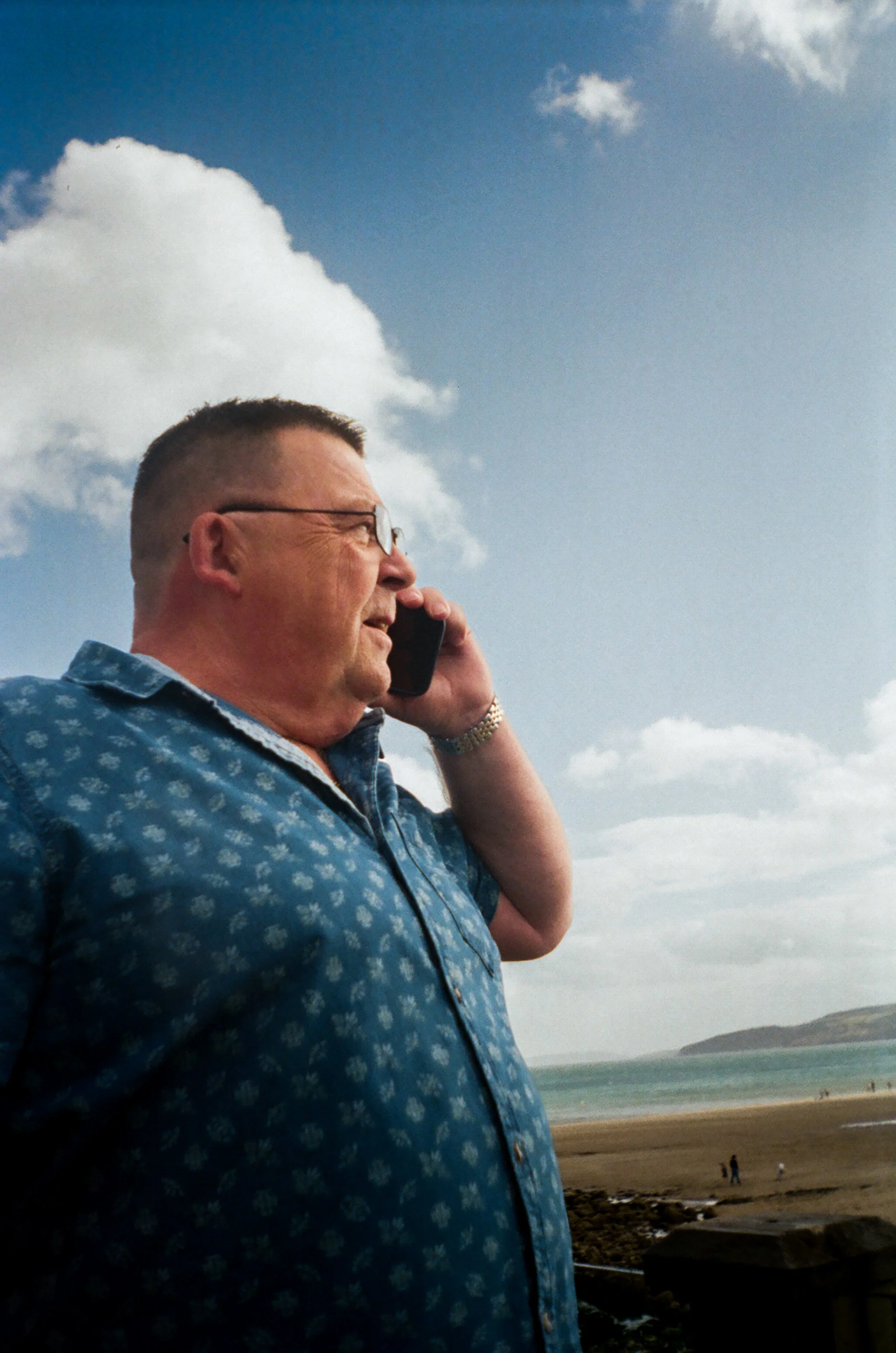 A man with glasses and a short haircut is talking on his cellphone at the beach during the daytime, with a cloudy sky and ocean in the background.