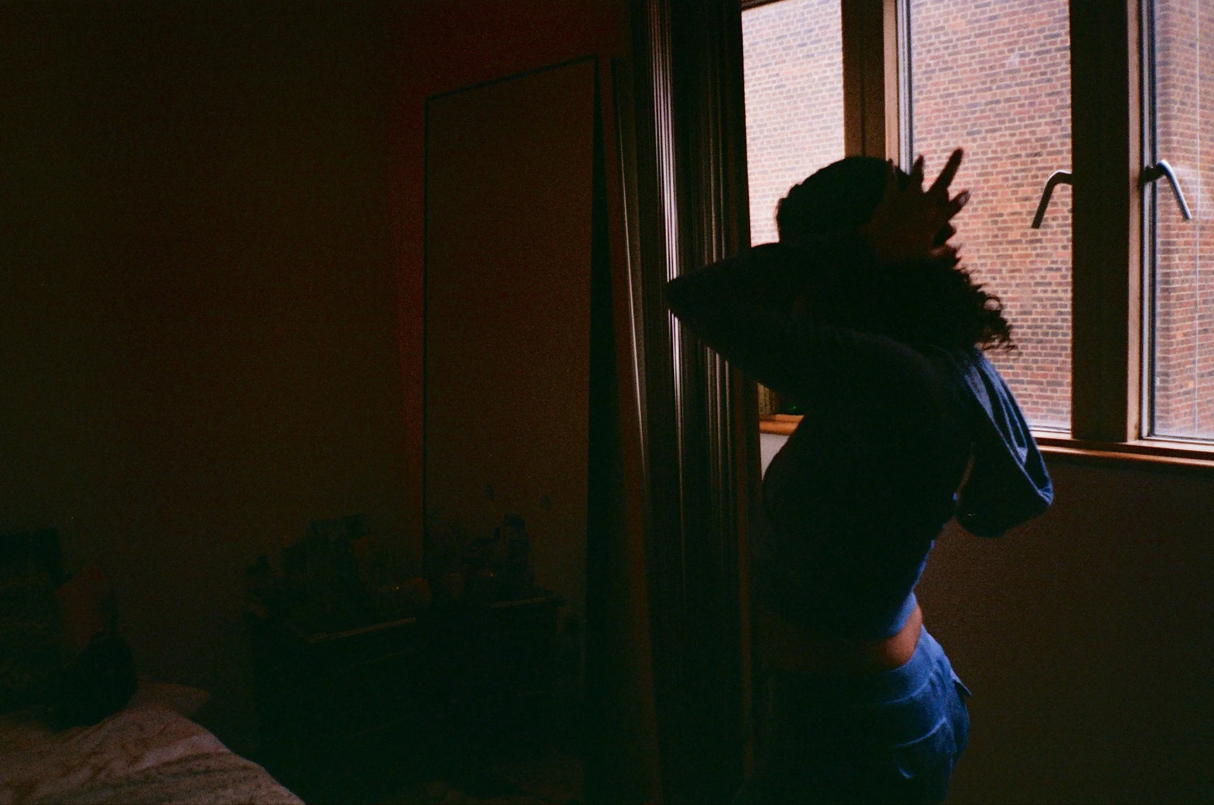 Person with curly hair standing in profile touching their head, inside a dimly lit room near a window.