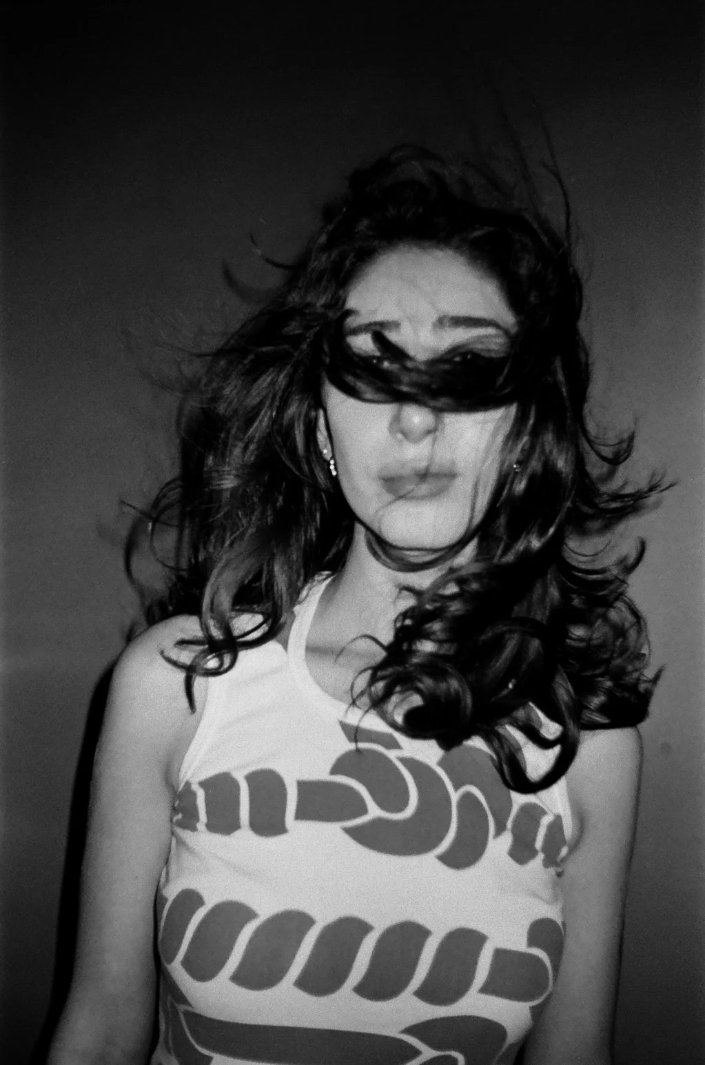Black and white photo of a woman with long wavy hair, partially covering her face with her hair, wearing earrings and a sleeveless t-shirt with a bold design.