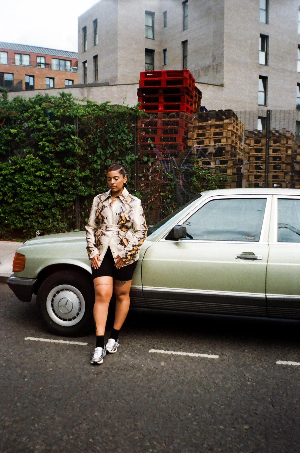 A young woman with dark hair and tattoos, wearing a snake print jacket, black shorts, and sneakers, stands beside a vintage green Mercedes-Benz car parked on the street. In the background, there are stacked pallets and urban buildings.