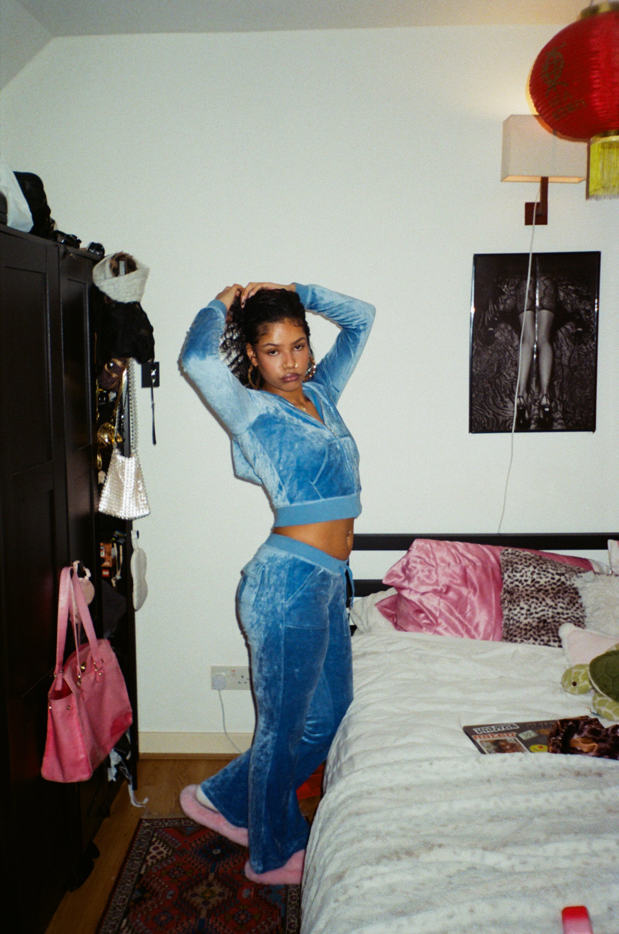 Young woman in a blue velour tracksuit standing in a bedroom, holding her curly hair with both hands, with pink slippers, a bed with pink and animal print pillows, and various items on a dark wood dresser nearby.