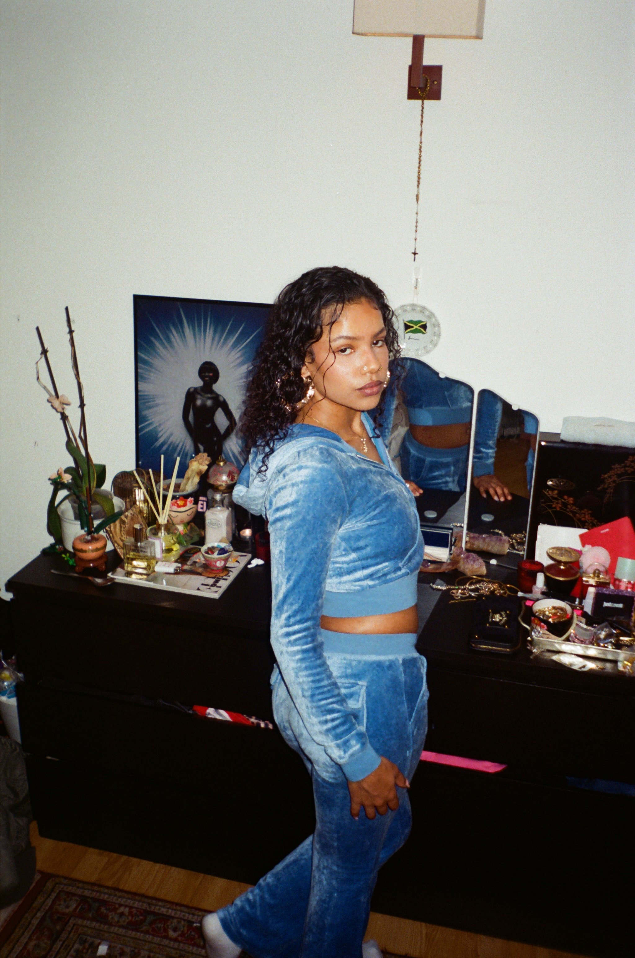 A young woman posing in a room with a dresser covered in various items, including jewelry, cosmetics, and decorative objects, with a mirror and a picture of a woman with a radiant background in the background.