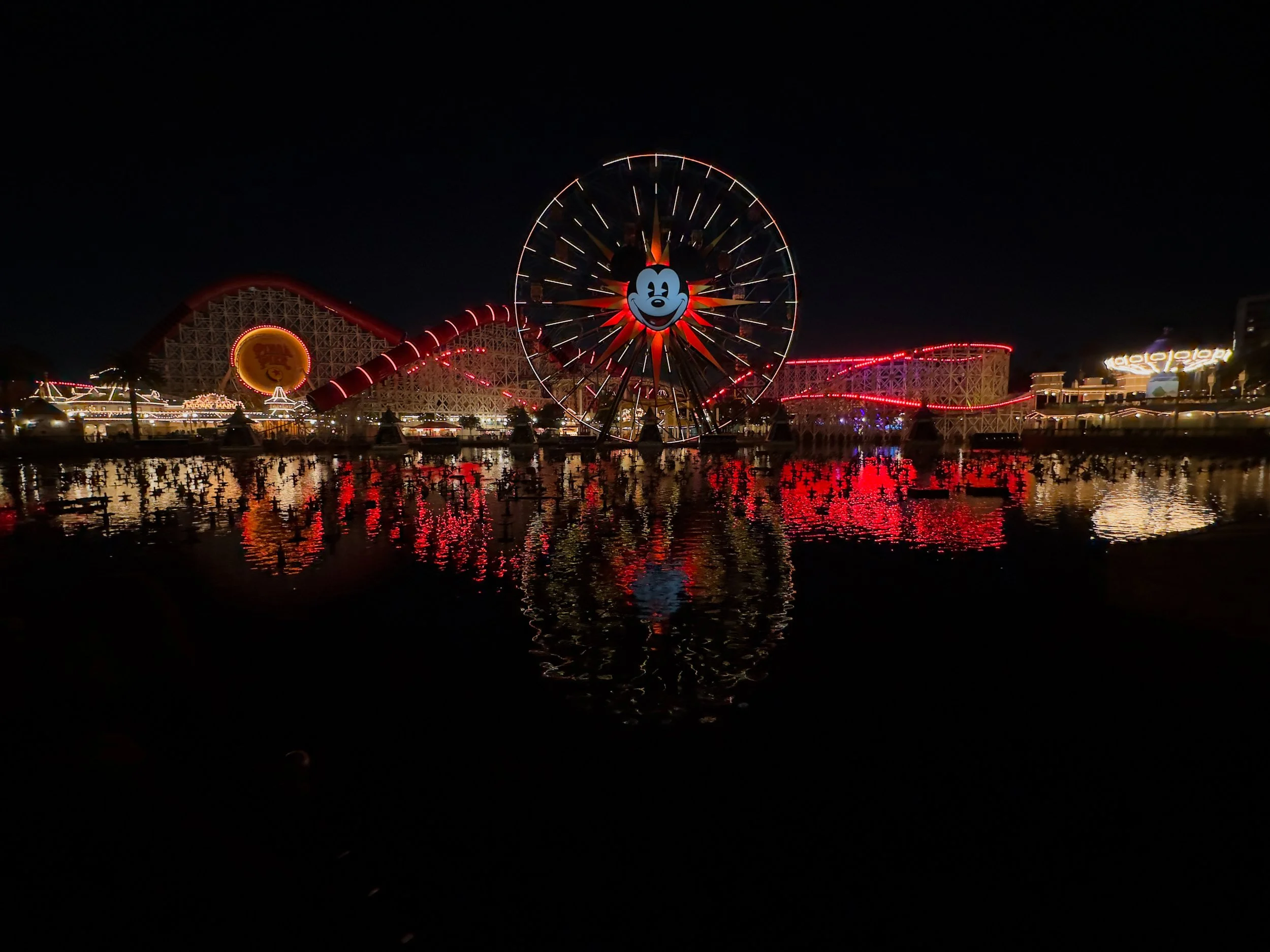 Nighttime view of an amusement park with a Mickey Mouse Ferris wheel illuminated in red and white, reflections on the water, and a wooden roller coaster with lights in the background.