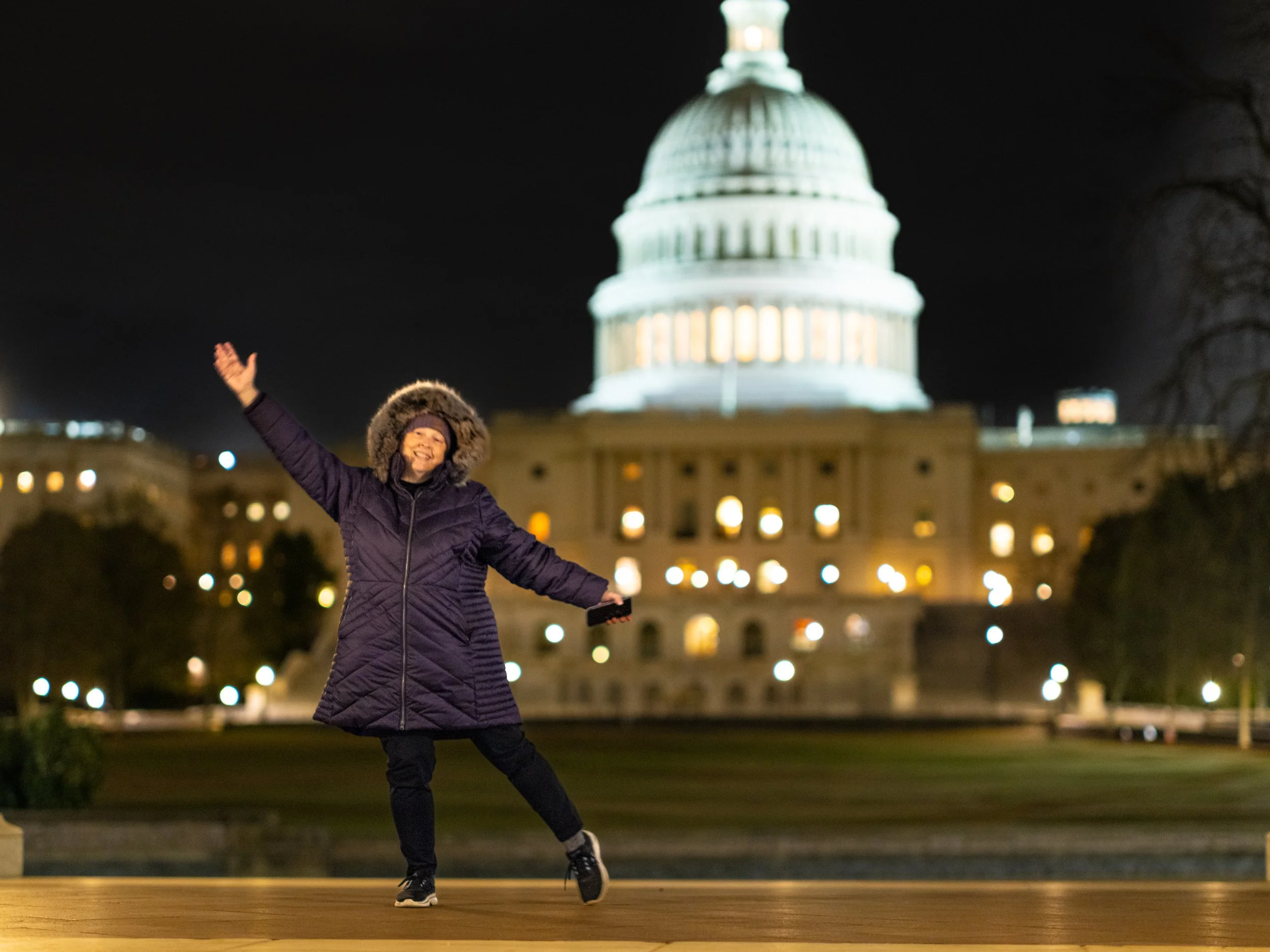 A Sheri Minchey Roberts wearing a purple winter coat with a fur-lined hood smiling and dancing outdoors at night in front of the illuminated United States Capitol building.