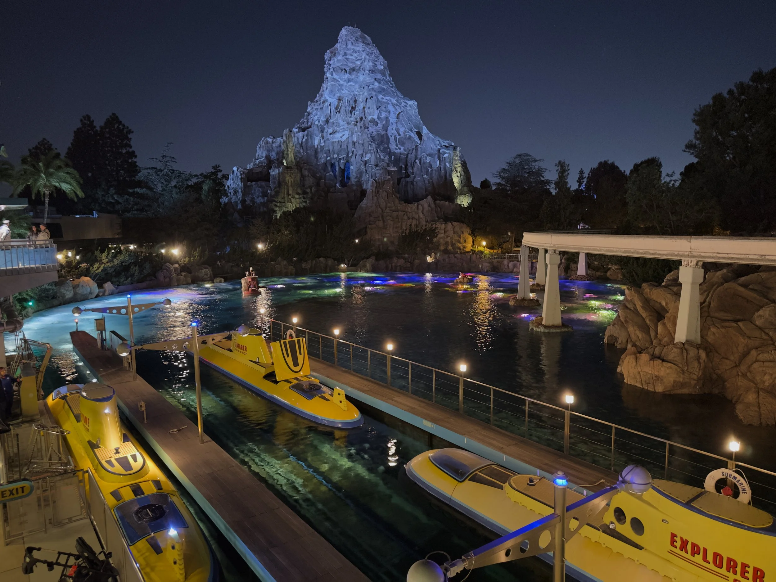 Night time view of in Disneyland of a boat ride with a large artificial mountain and various amusement rides, including yellow submarines and colorful lighting in the water.