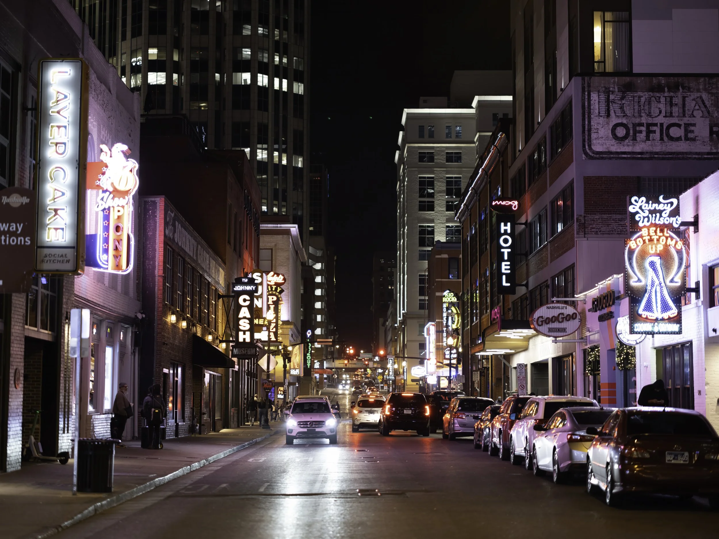 Night city street filled with parked cars and illuminated neon signs, including a large flashing sign for LAVE RACK and other colorful advertisements, with tall buildings in the background.