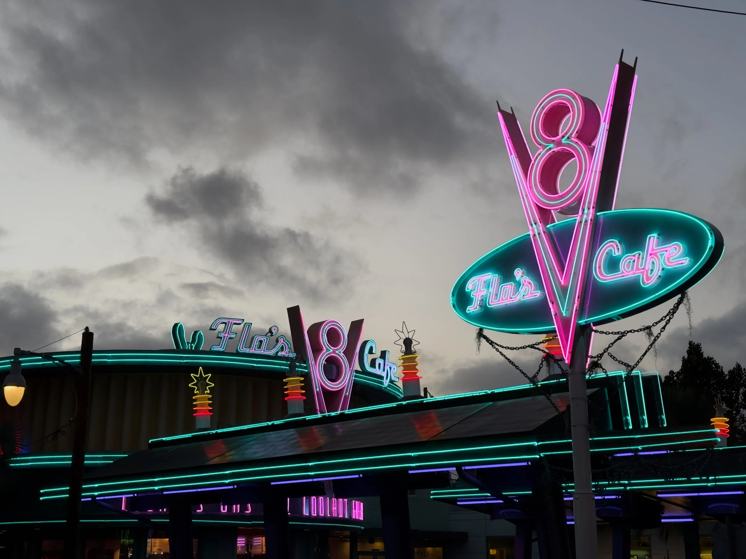Neon sign for a dessert shop called 'Flo's V-8 Cafe' with the words 'Fresh Flo's Cafe' illuminated in pink and blue lights, against a cloudy sky.