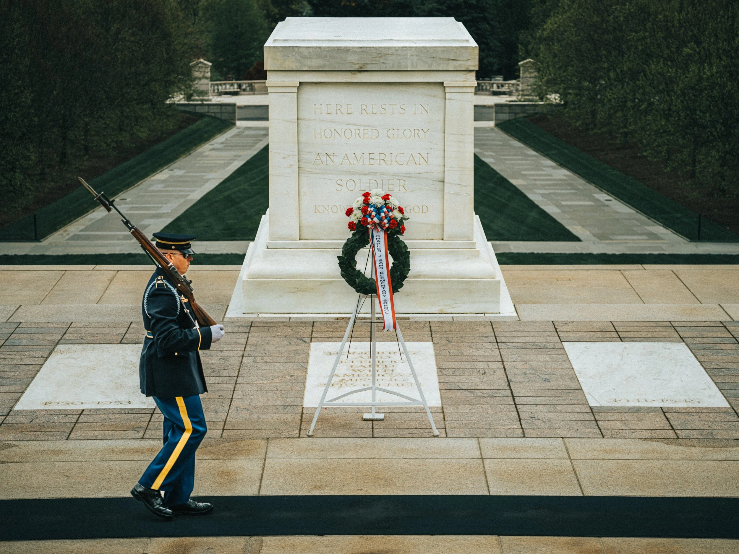 A soldier in a formal uniform standing guard with a rifle at the Tomb of the Unknown Soldier in Arlington National Cemetery. The tomb has an inscription that reads, 'Here Rests in Honored Glory an American Soldier.' There is a wreath with red, white,