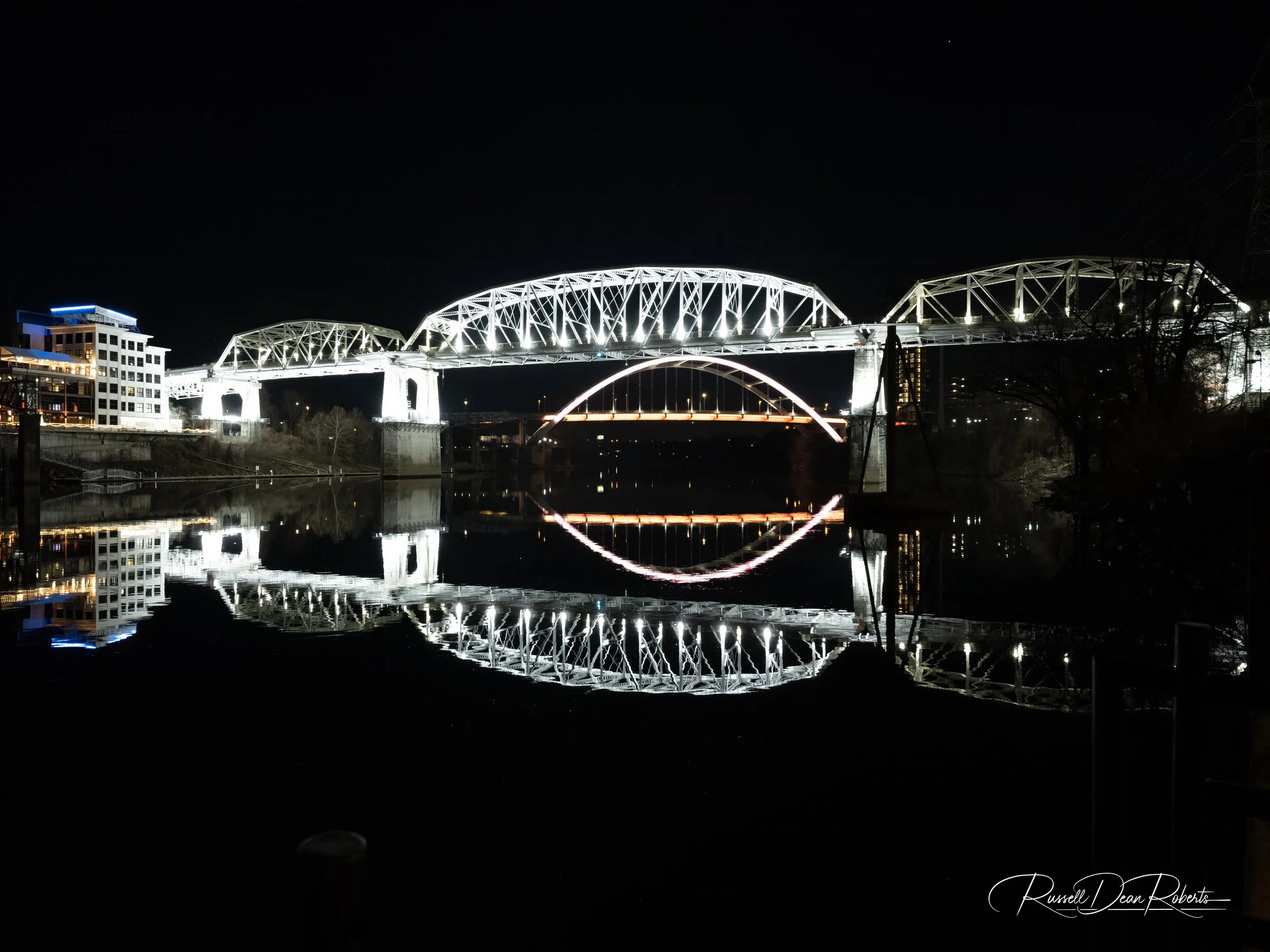 Nighttime cityscape in Nashville, Tennessee with the John Seigenthaler Pedestrian Bridge and its reflection on water, with buildings and trees nearby.