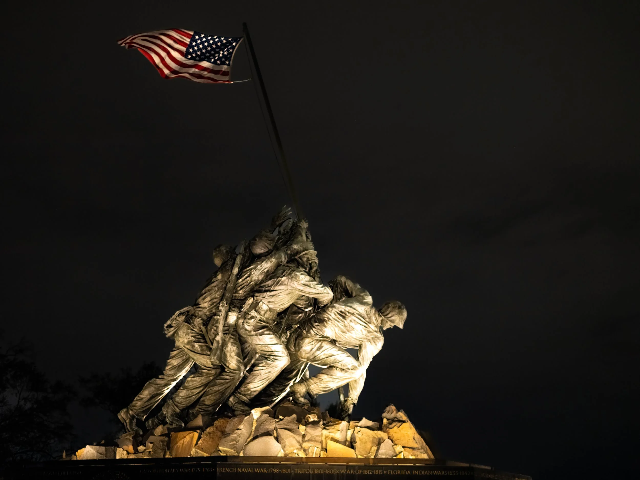 Nighttime view of the Marine Corps War Memorial featuring a sculpture of soldiers raising the American flag, illuminated against a dark sky, with a rocky base at the bottom.
