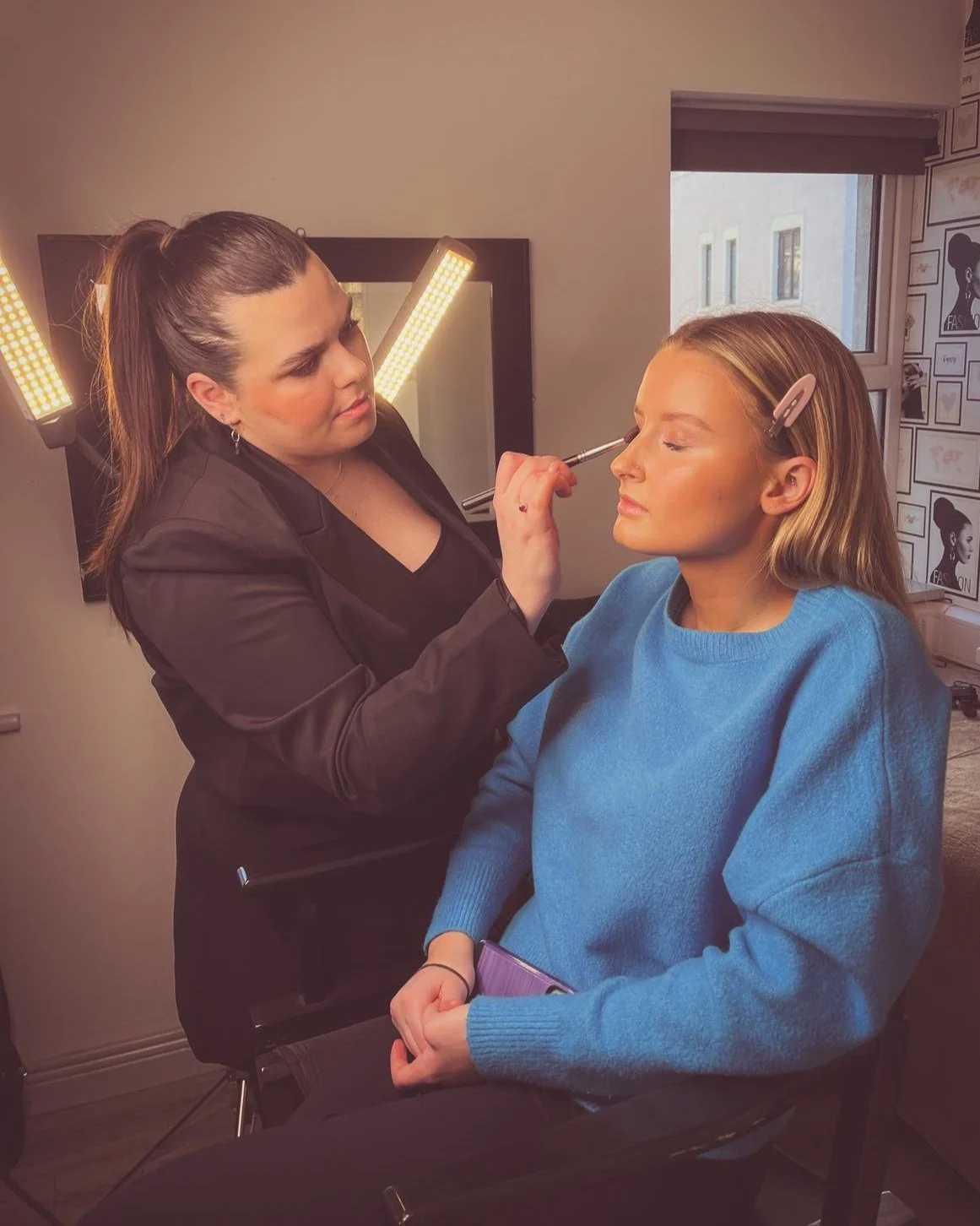 Makeup artist applying makeup to woman sitting in a chair, with a mirror and studio lights in the background.