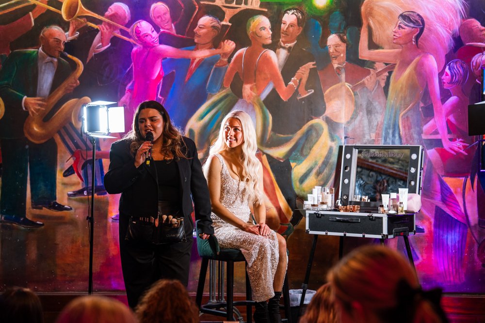 A woman with long blonde hair in a sparkly dress is seated on a chair on stage, smiling at a woman standing next to her with a microphone. The stage features colorful mural art of jazz musicians and dancers, with bright lighting and audience members visible in the foreground.