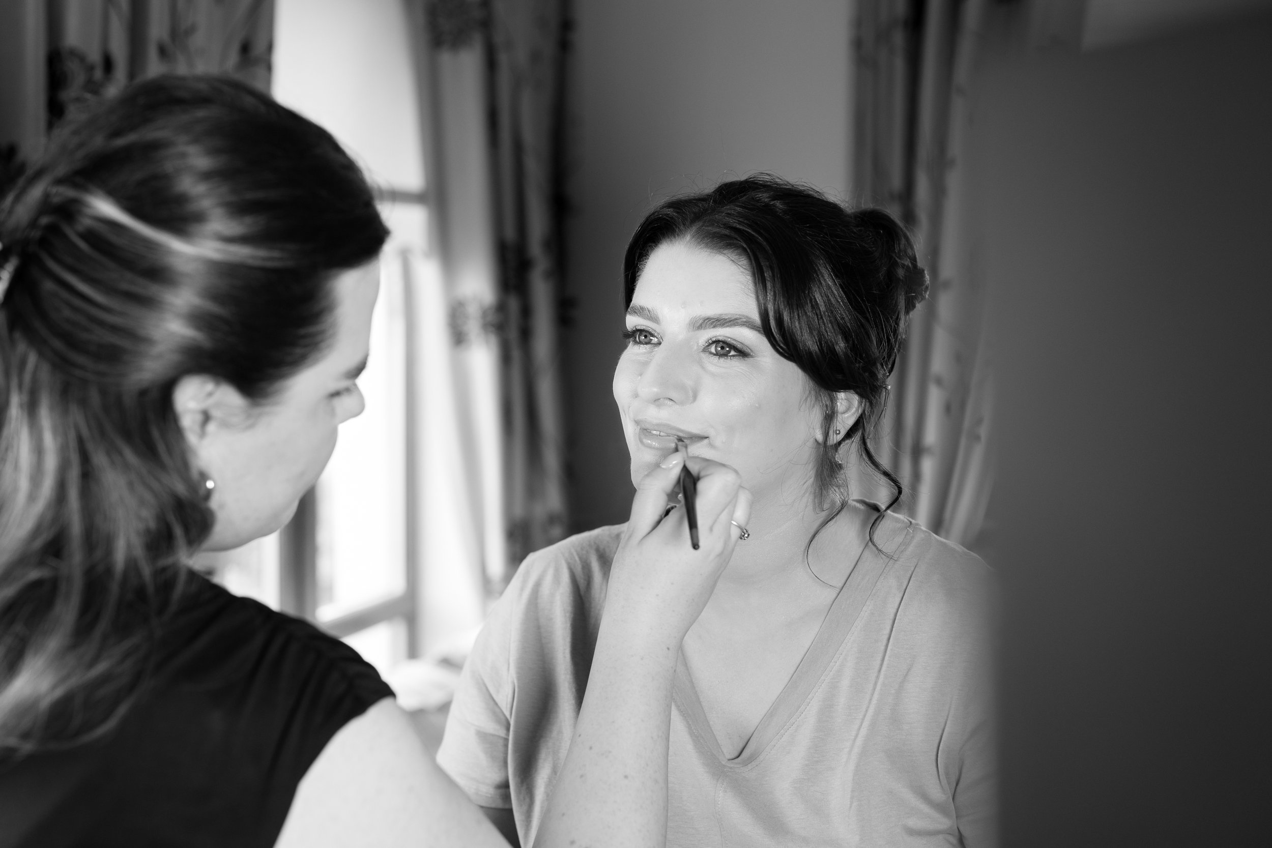 A woman with dark hair and light skin getting her makeup done by a makeup artist in a room with curtains and a window.