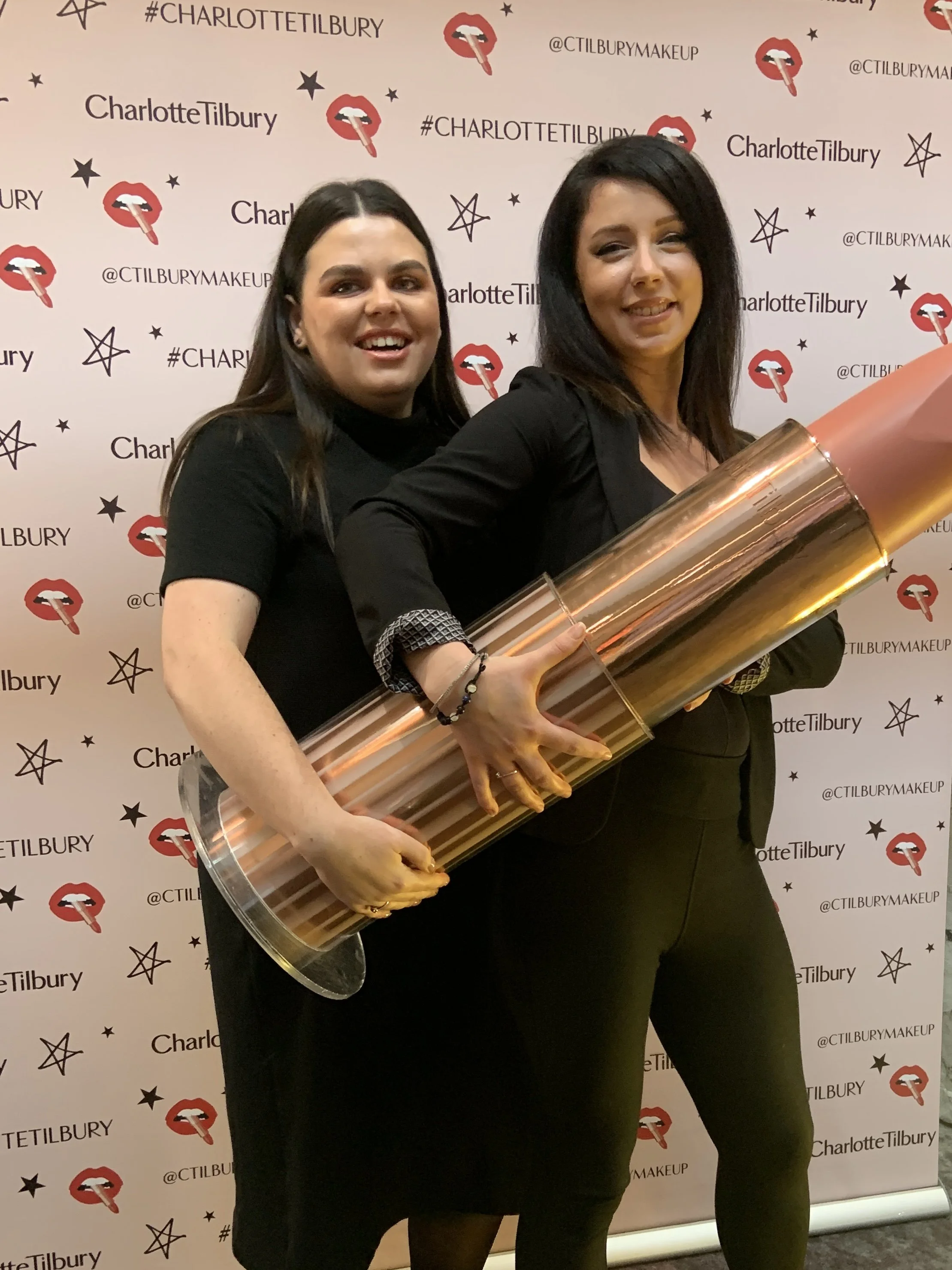 Two women are standing in front of a Charlotte Tilbury branded backdrop, smiling and holding a large lipstick prop.