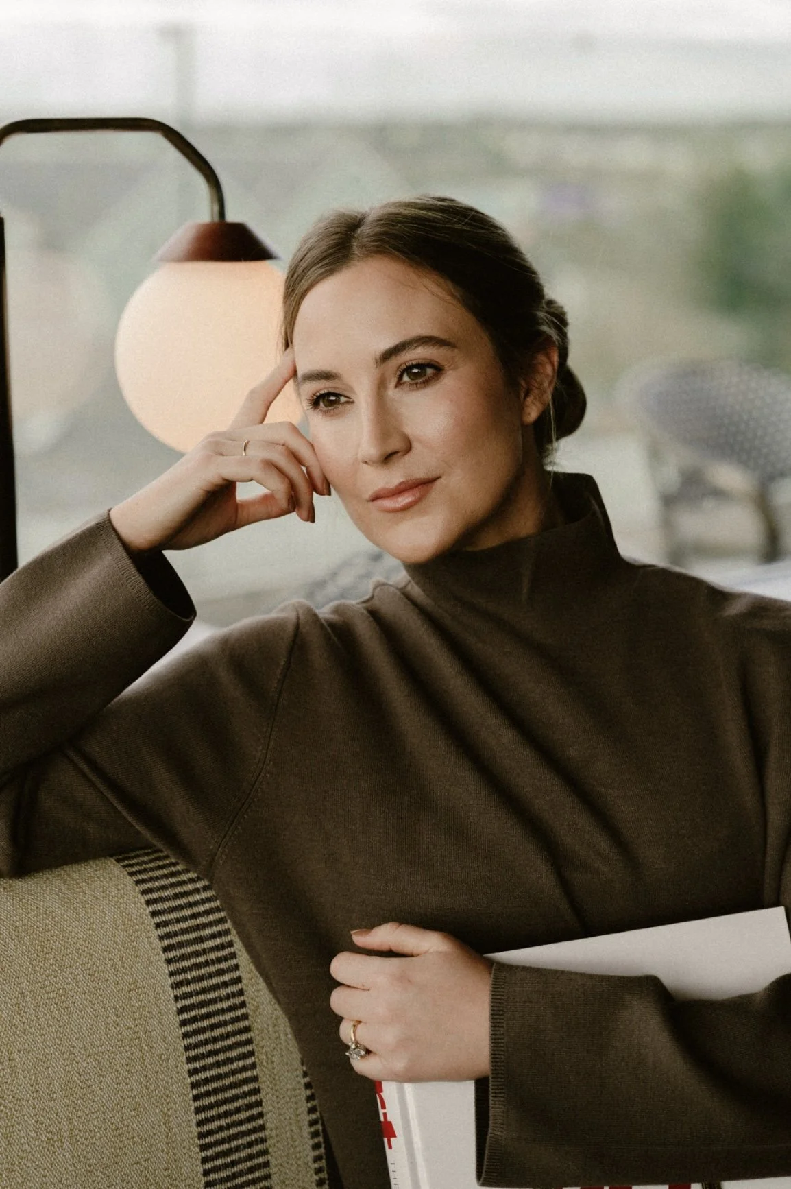A woman with brown hair styled in a low bun is sitting indoors, resting her head on her left hand, with her eyes slightly to the side and a thoughtful expression. She is wearing a brown turtleneck sweater and holding a white book or folder under her right arm, which rests on a yellow and black-striped chair. There is a round hanging lamp and a large window in the background.