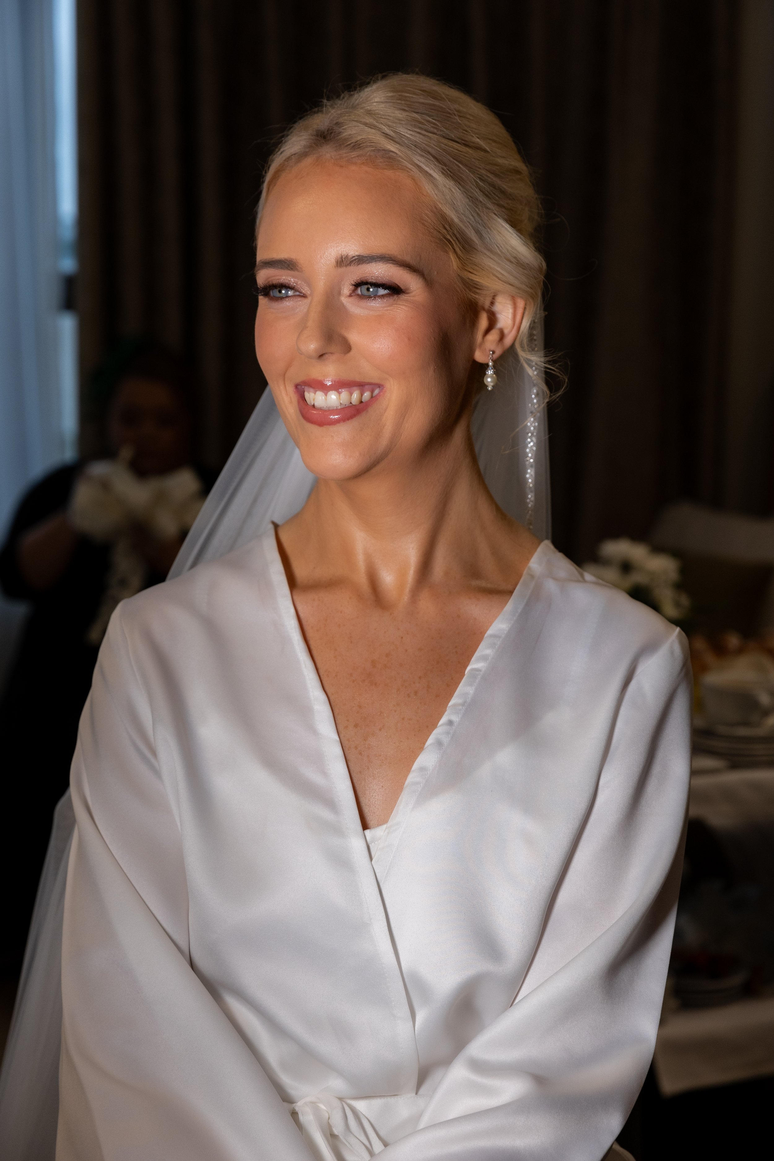 A smiling bride with blonde hair styled in an updo, wearing pearl earrings and a white satin robe, getting ready for her wedding.