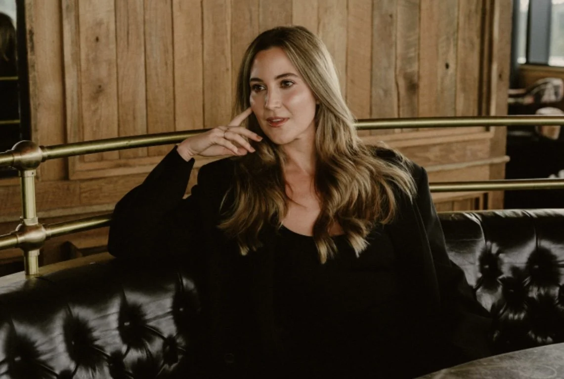 A woman with long wavy blonde hair sitting on a black leather tufted sofa in a wooden-paneled room.
