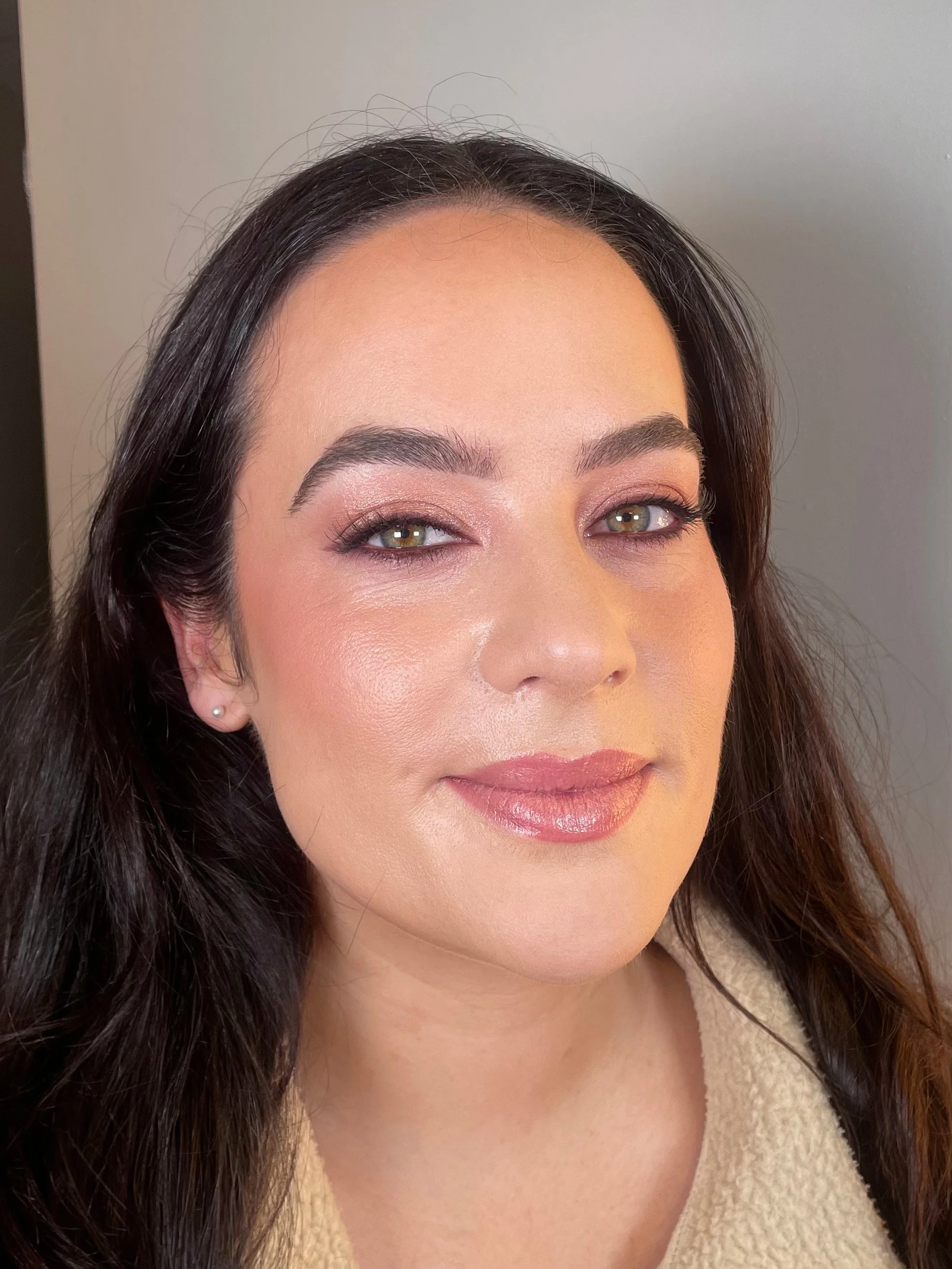 Close-up of a woman with dark hair, green eyes, and makeup, wearing a beige top and small pearl earrings, smiling softly in front of a plain gray background.