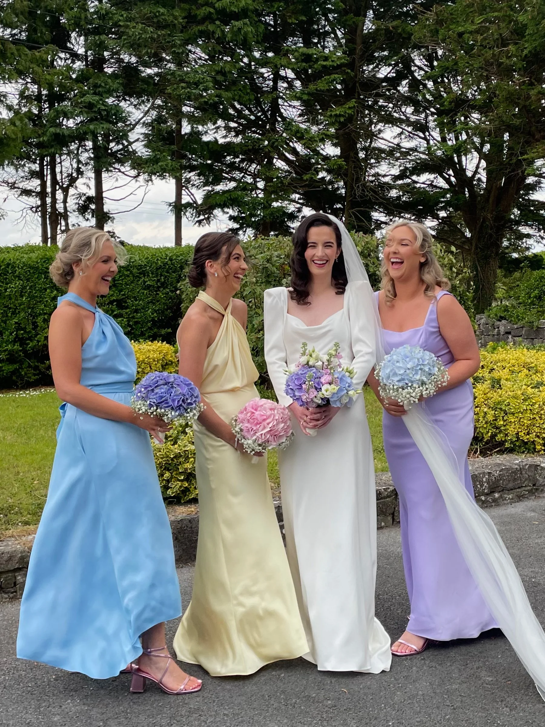 A bride in a white wedding gown with a veil stands surrounded by three bridesmaids in pastel-colored dresses outdoors, all holding bouquets and laughing.