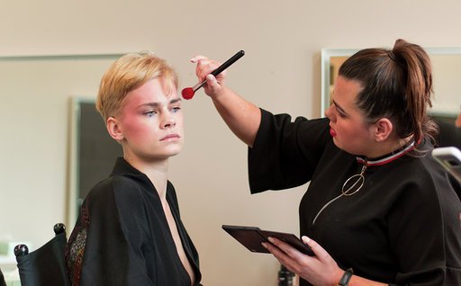 A woman applying makeup with a brush to a young person's face in a makeup chair in a room with neutral-colored walls.