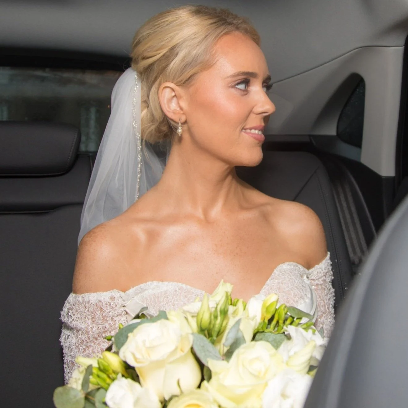 A bride with blonde hair sitting in a vehicle, holding a bouquet of white and cream flowers, wearing pearl earrings and an off-the-shoulder wedding dress with lace details.