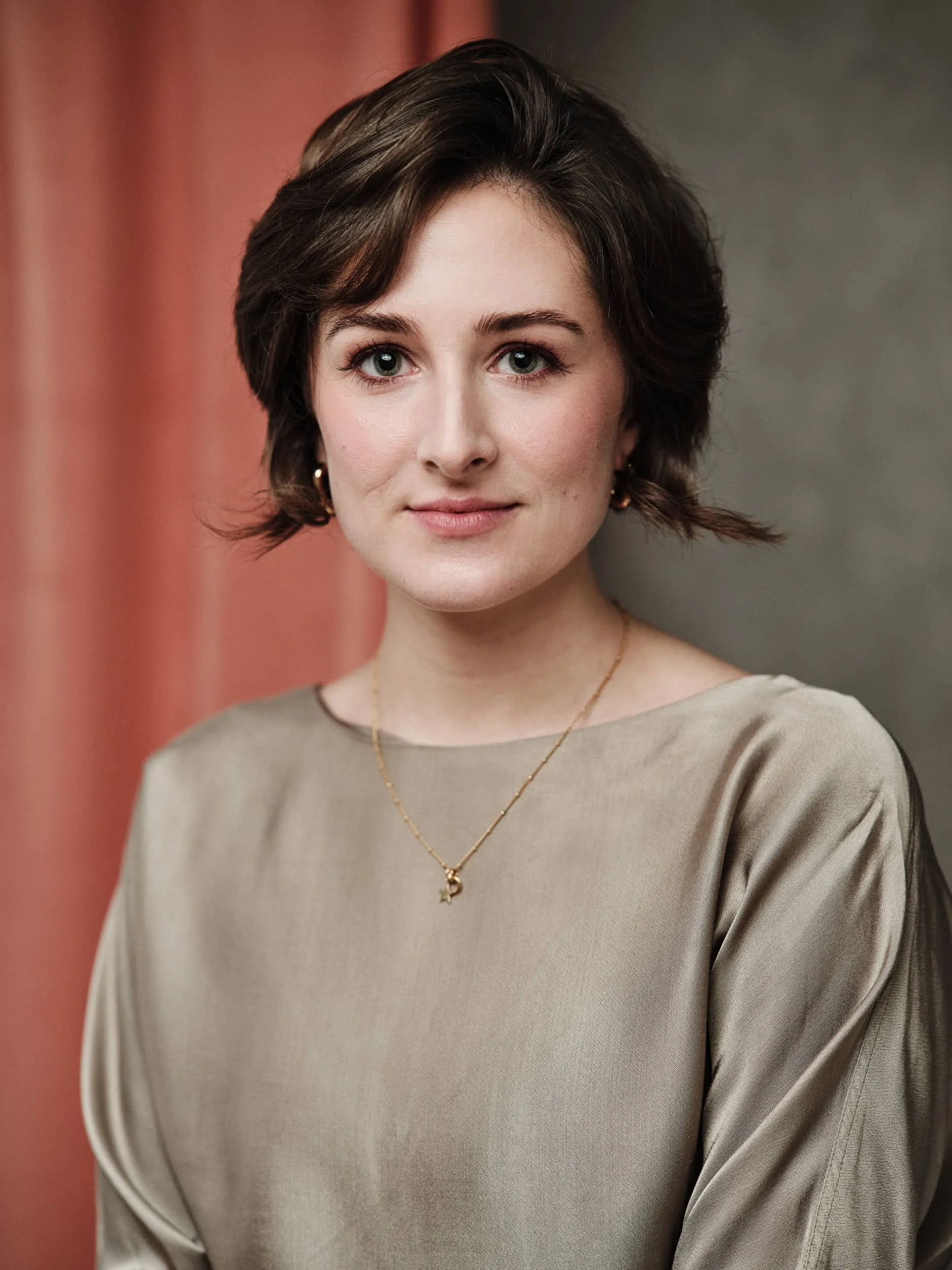 Portrait of a woman with short brown hair, wearing a beige satin blouse and gold jewelry, standing in front of a background with muted pink and grey tones.