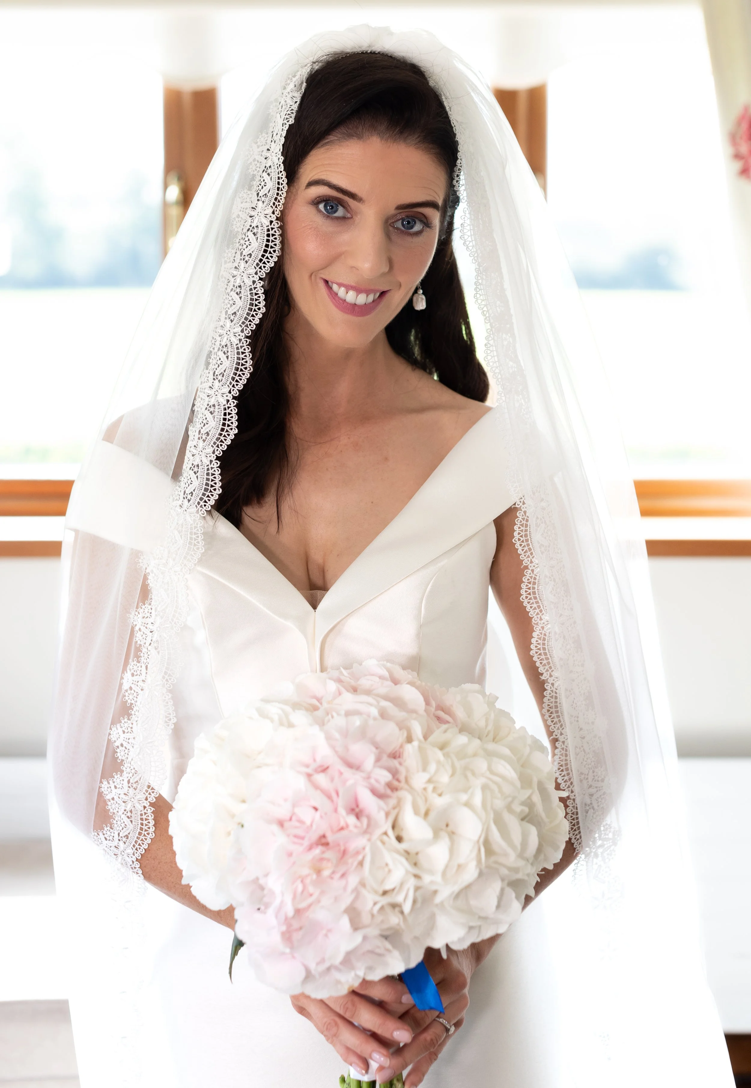 A bride with dark hair and blue eyes wearing a white wedding dress and veil, holding a bouquet of white and pink flowers, smiling indoors.