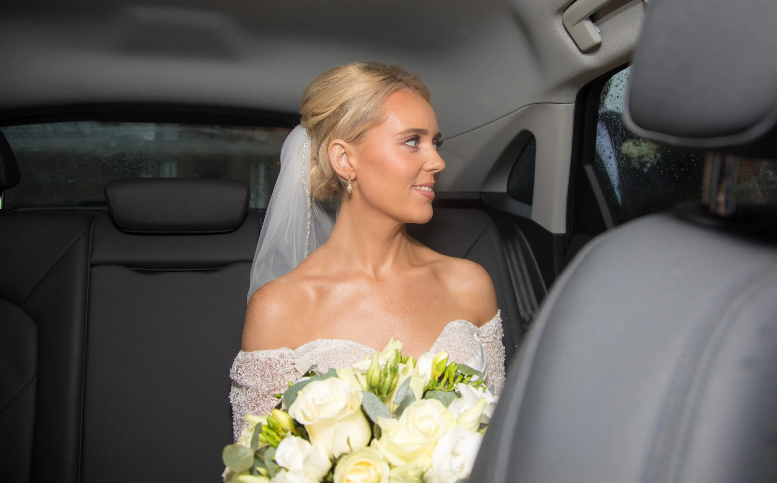 Bride in wedding dress and veil sitting in the backseat of a car, holding a bouquet of white roses, looking to her right.