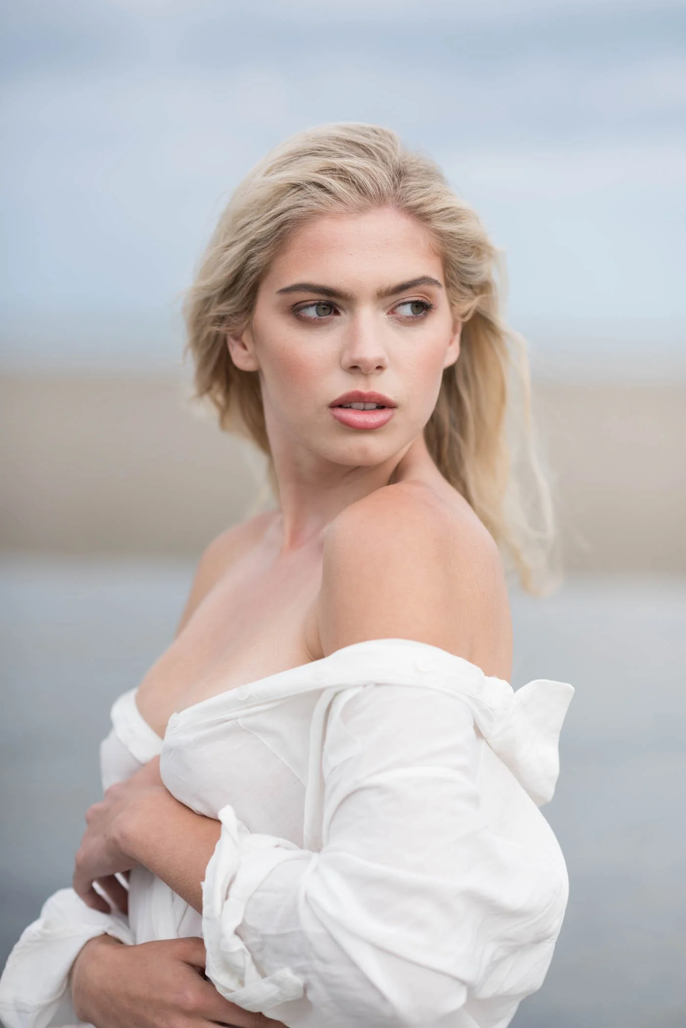 A blonde woman with shoulder-length hair and fair skin is looking to her right. She is wearing a white off-the-shoulder shirt that is slightly slipping off one shoulder, and she is holding the shirt with her left hand. The background appears to be an