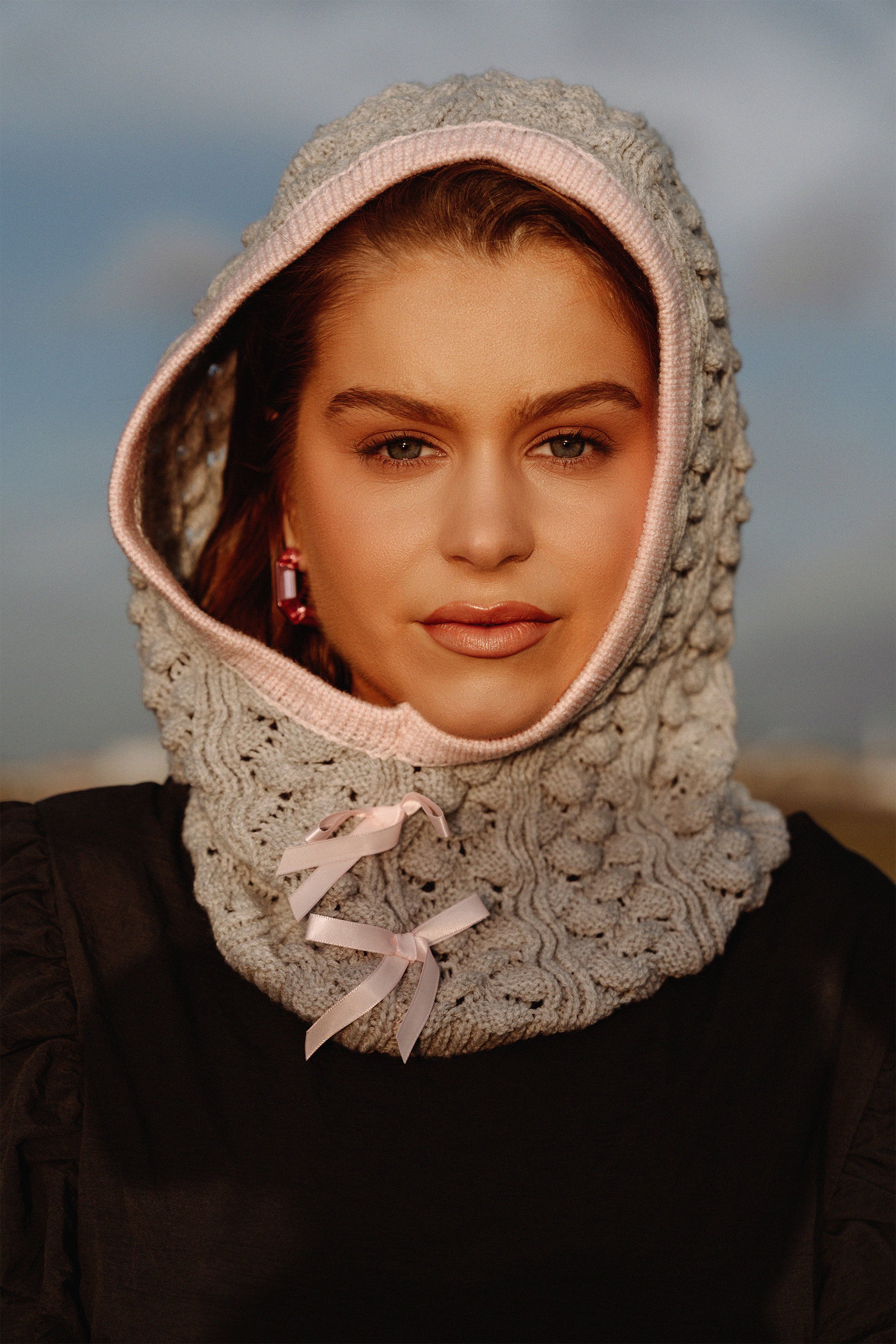 A young woman wearing a knitted gray hood with pink trim and ribbon ties, posing outdoors during sunset.