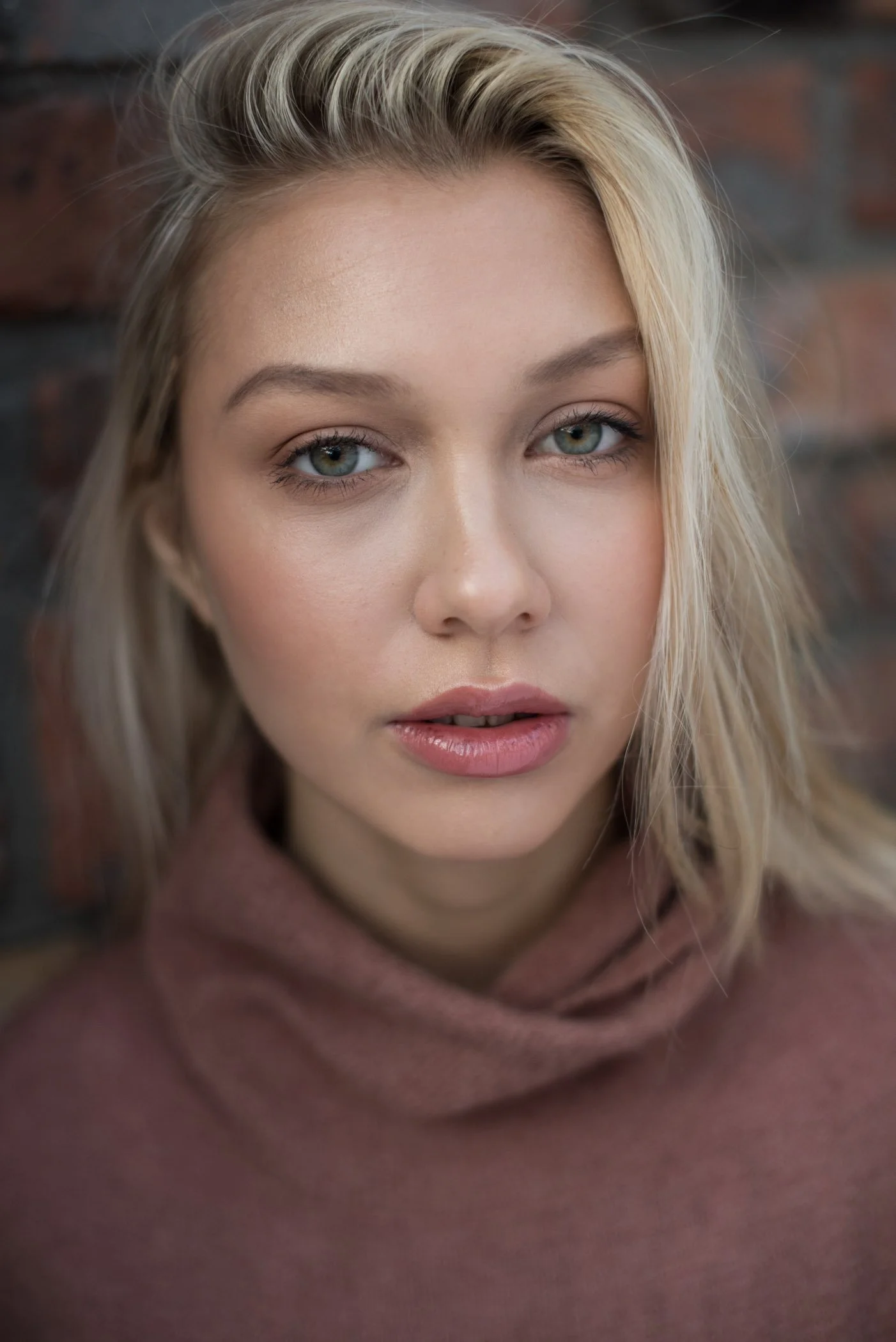 Close-up of a young woman with blonde hair, blue eyes, and a neutral expression, wearing a pinkish-brown sweater, against a brick wall background.