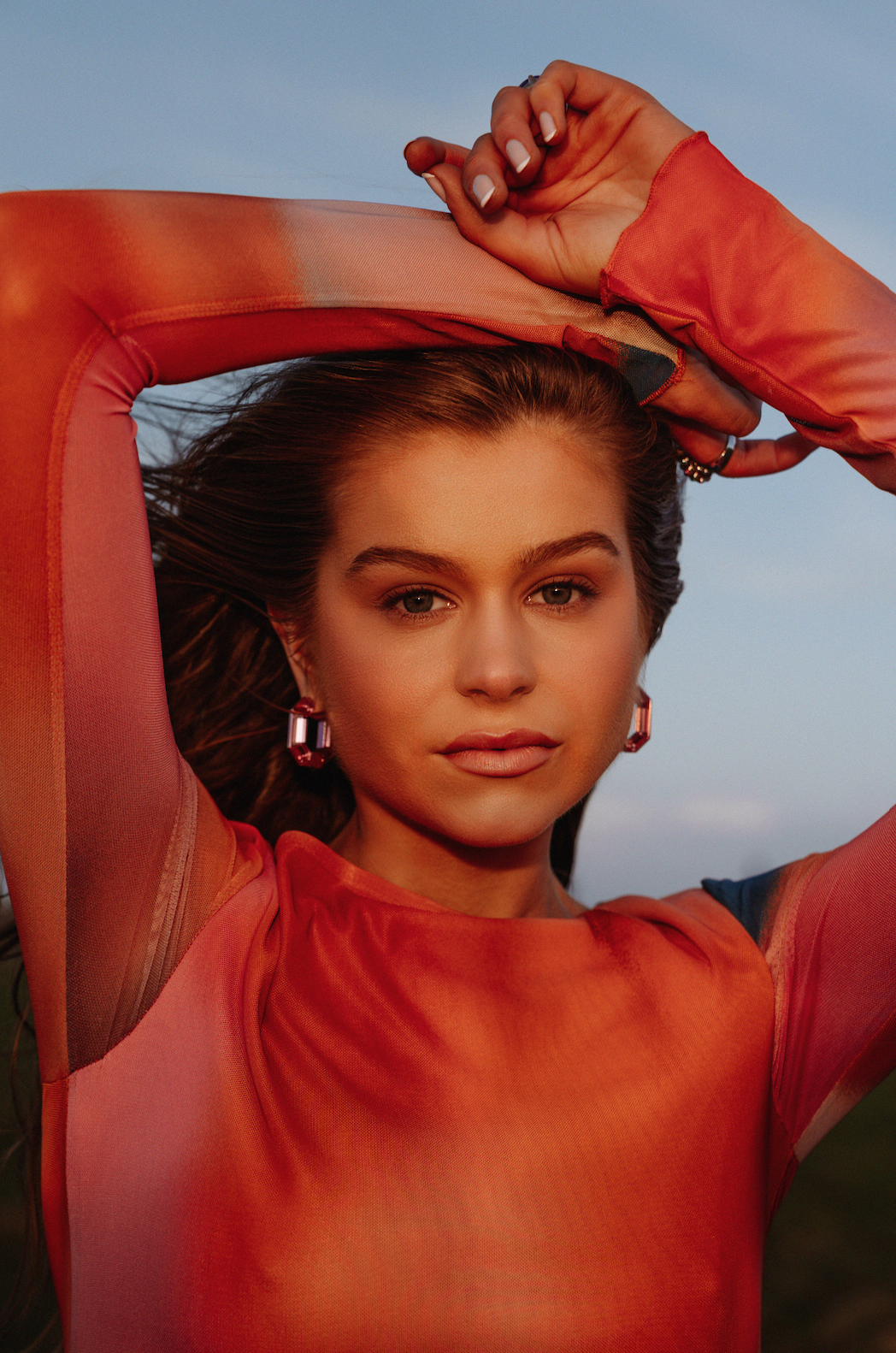 A woman with long hair and earrings wearing an orange and pink top, posing outdoors with arms raised and hands clasped above her head, during sunset.