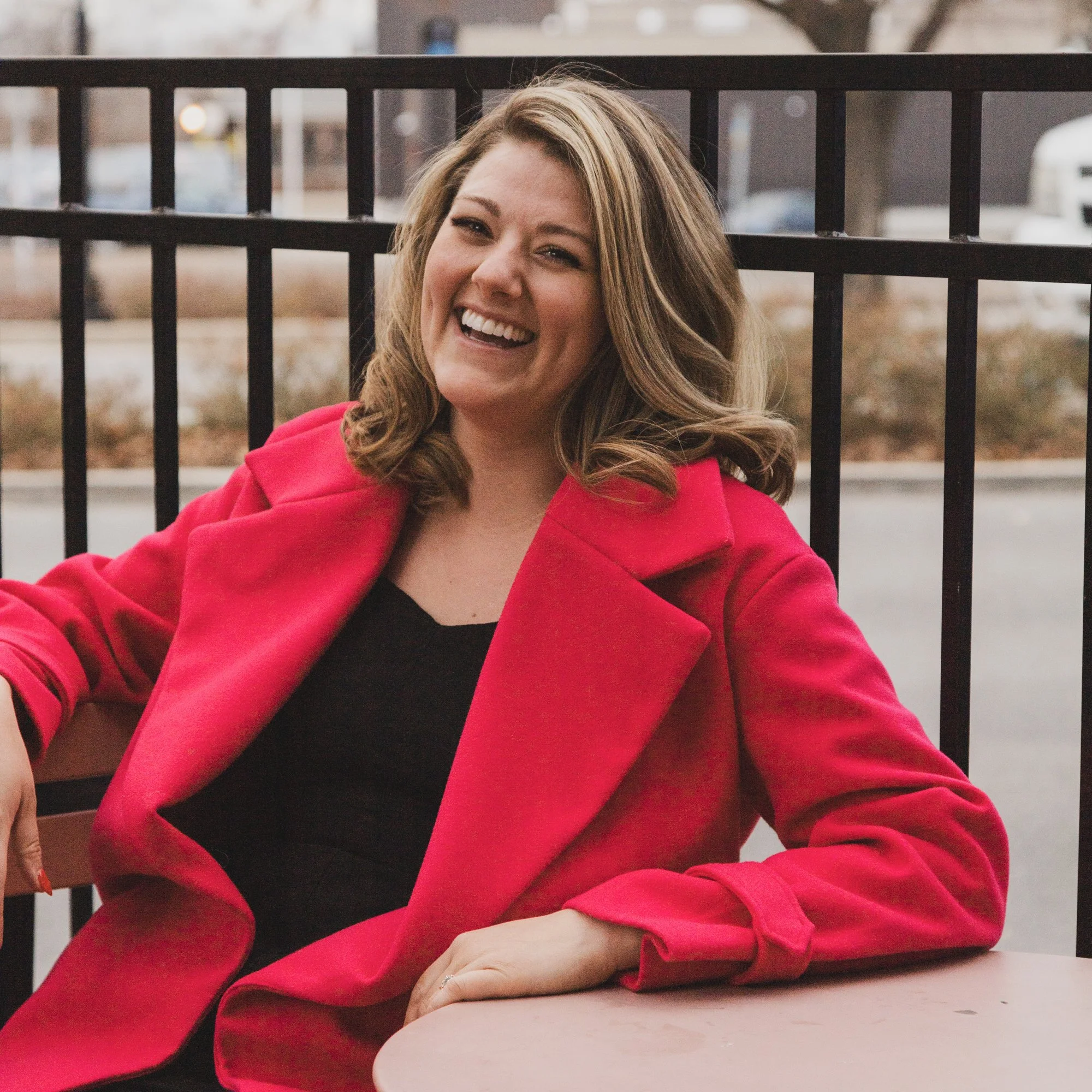 A woman with shoulder-length light brown hair, smiling, wearing a red coat and black top, sitting outdoors at a table with a black railing behind her.