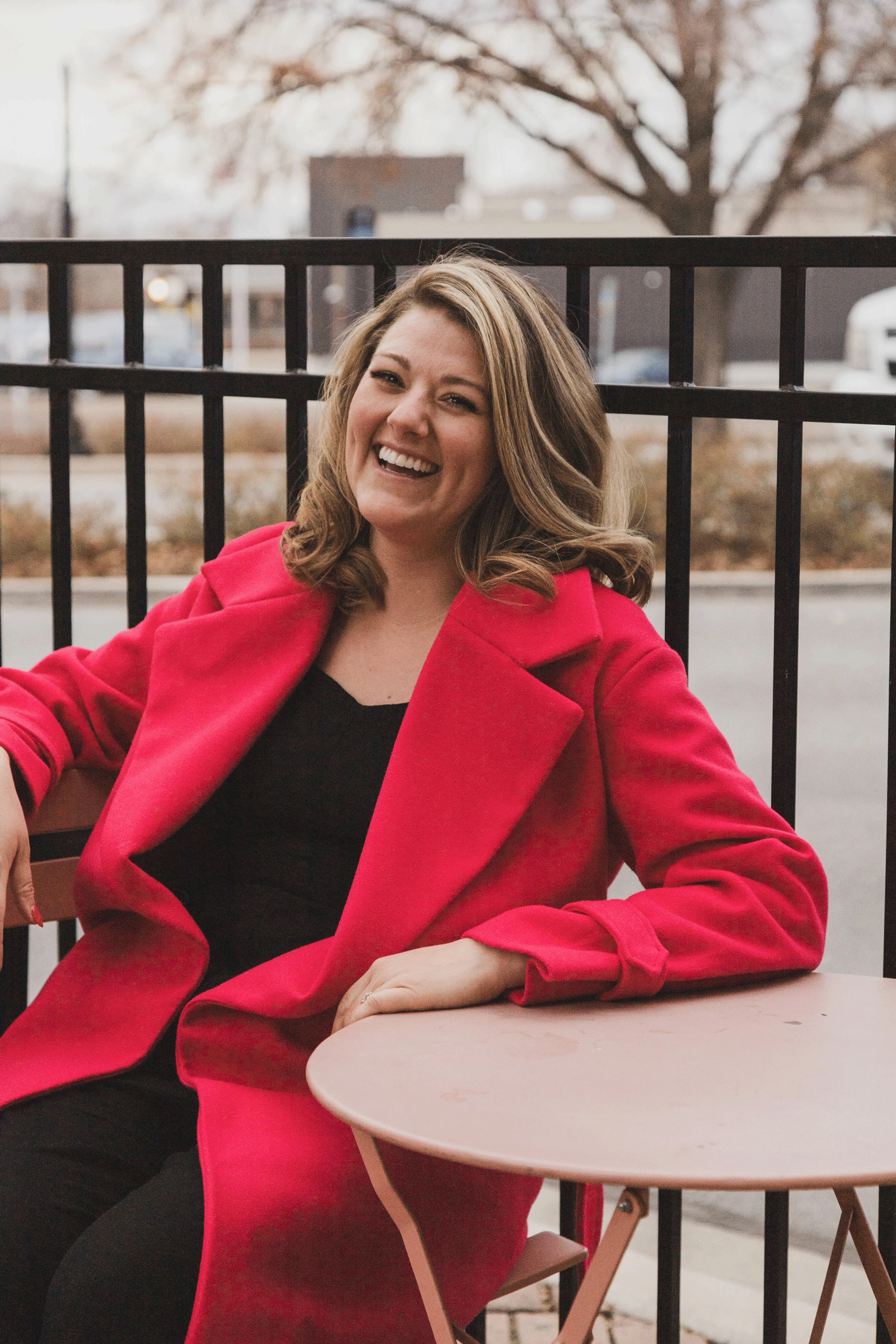 A woman with shoulder-length brown hair, smiling and wearing a bright red coat, sitting outdoors at a small round pink table.