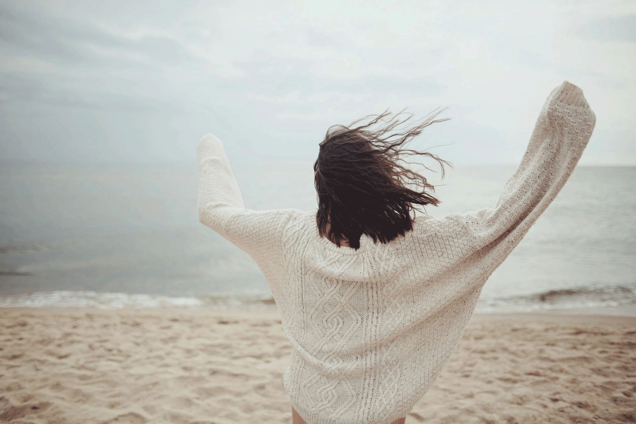A woman with dark hair in a beige sweater standing on a sandy beach with her arms outstretched, facing the water and sky, windy day.