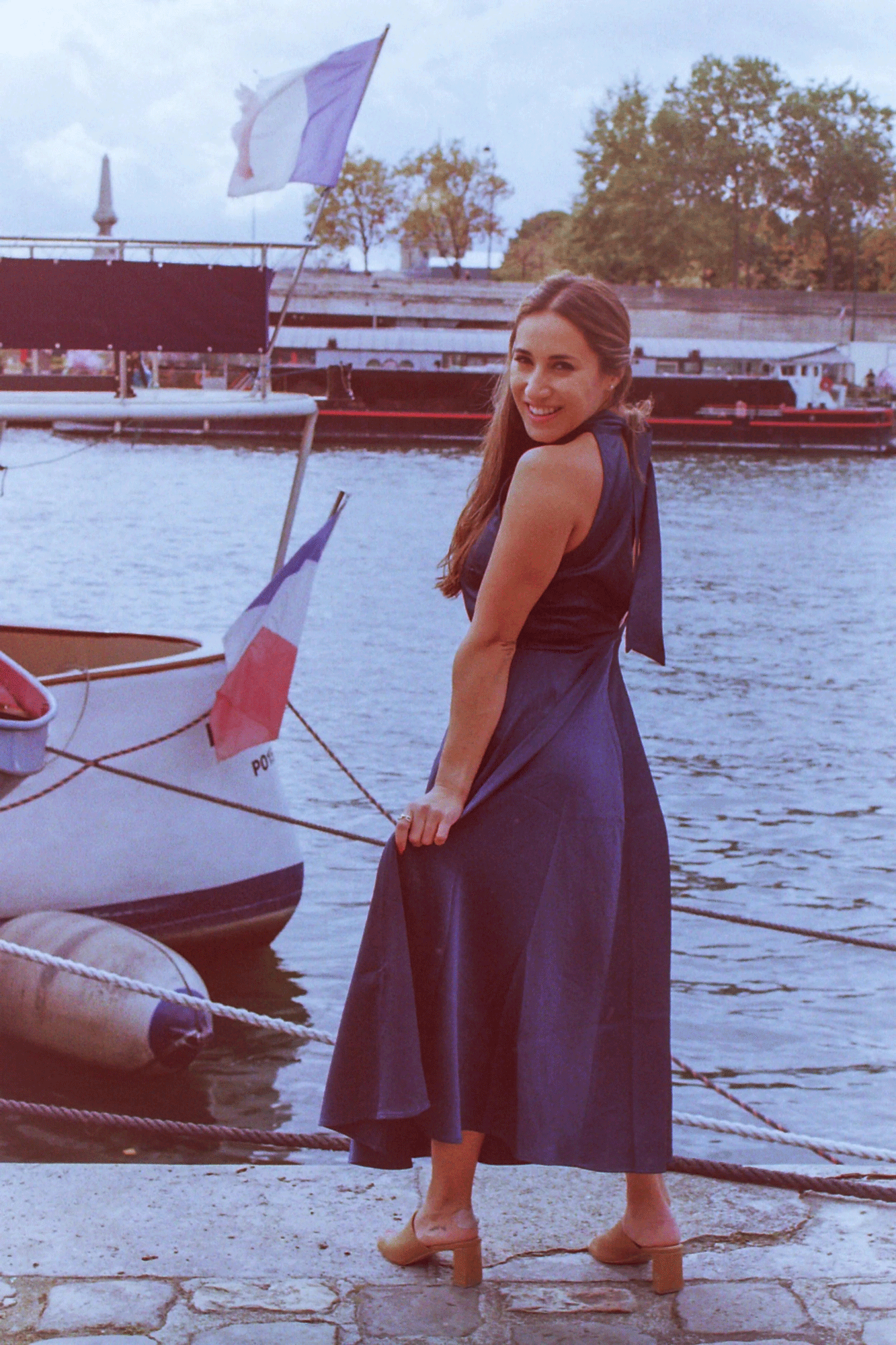 A woman in a blue dress standing by a harbor with boats and French flags, smiling at the camera.