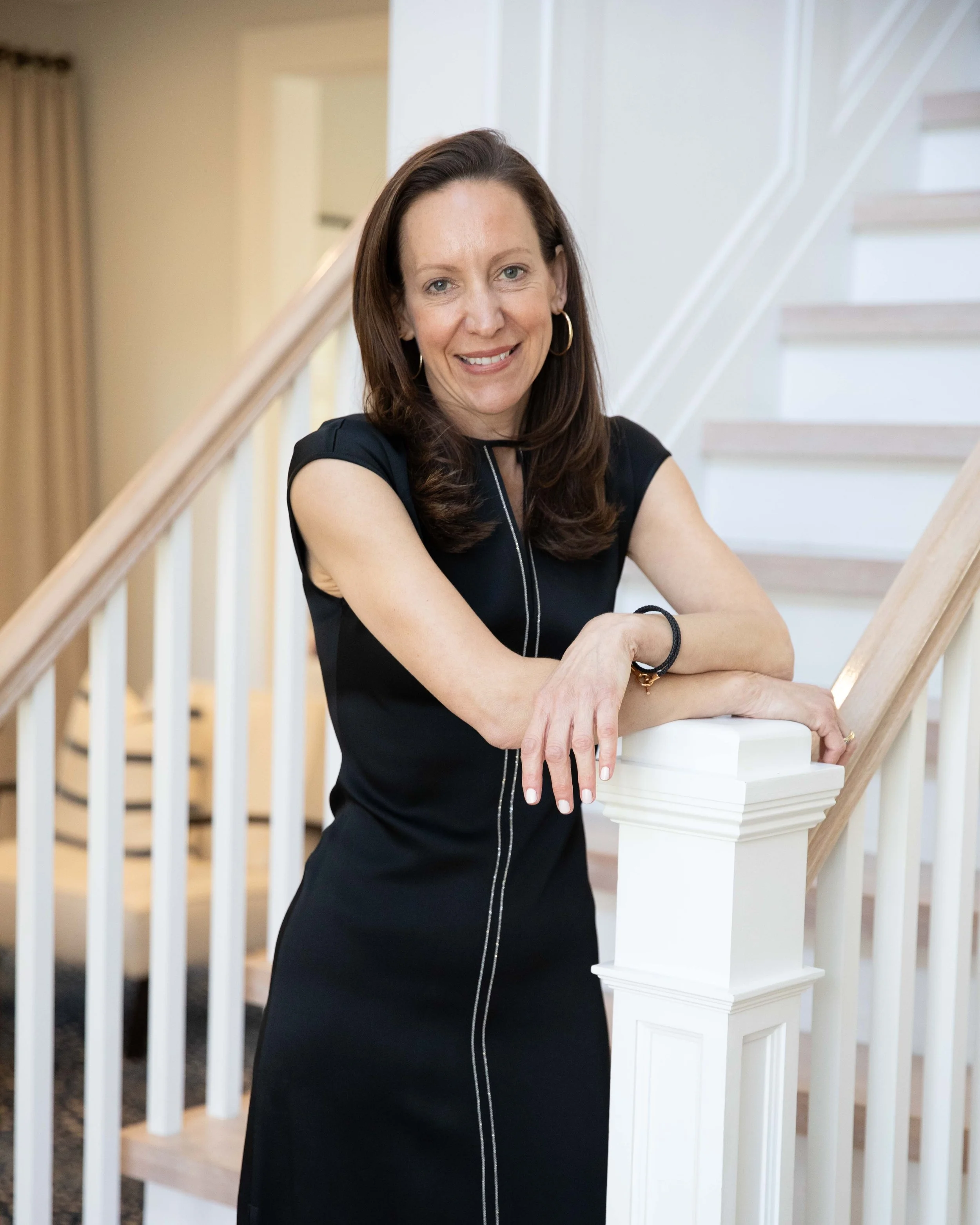 A woman with brown hair, wearing a black dress, leaning on a staircase railing and smiling.