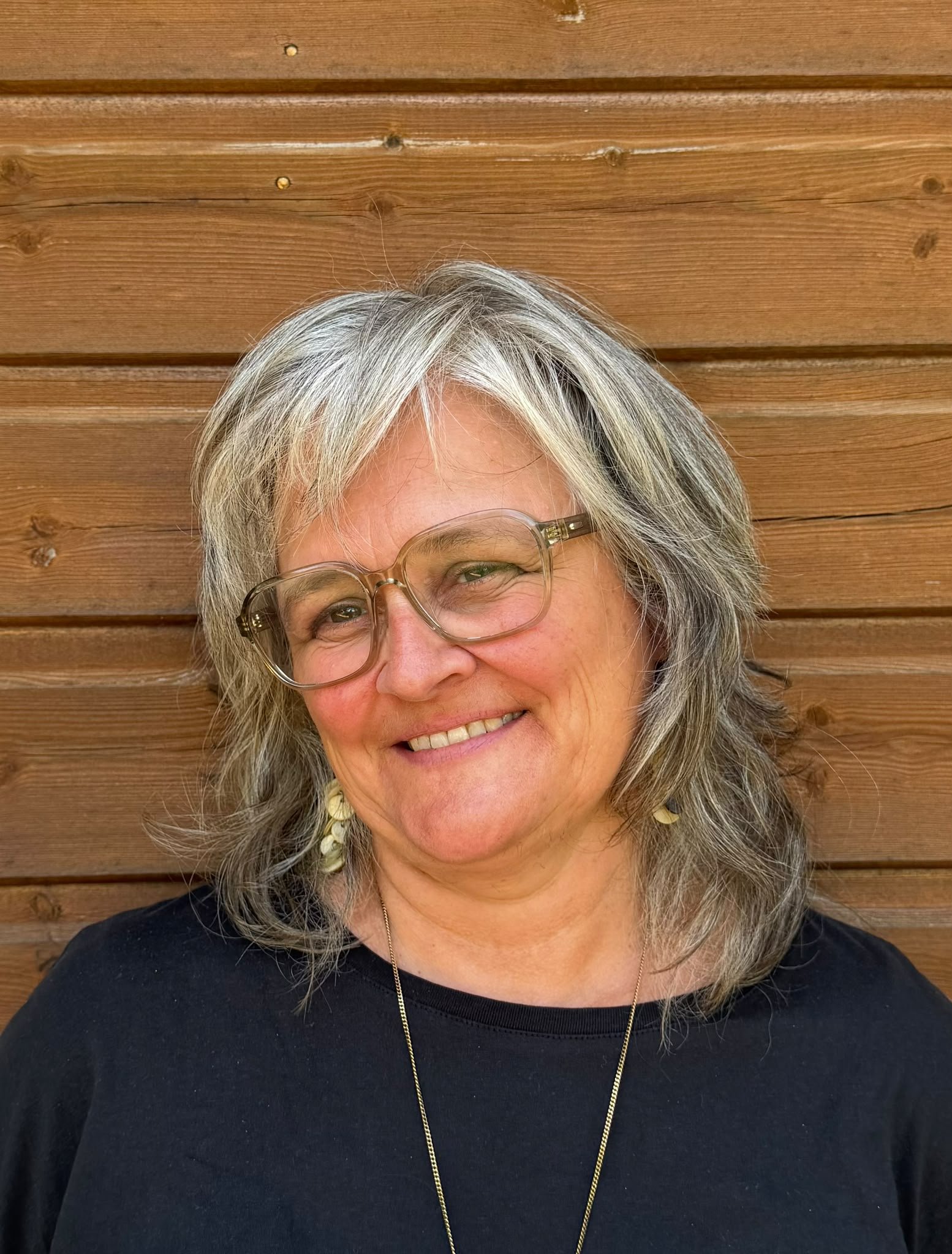 Close-up photo of a smiling woman with gray hair, wearing glasses, a black top, and gold jewelry, standing in front of a wooden wall.