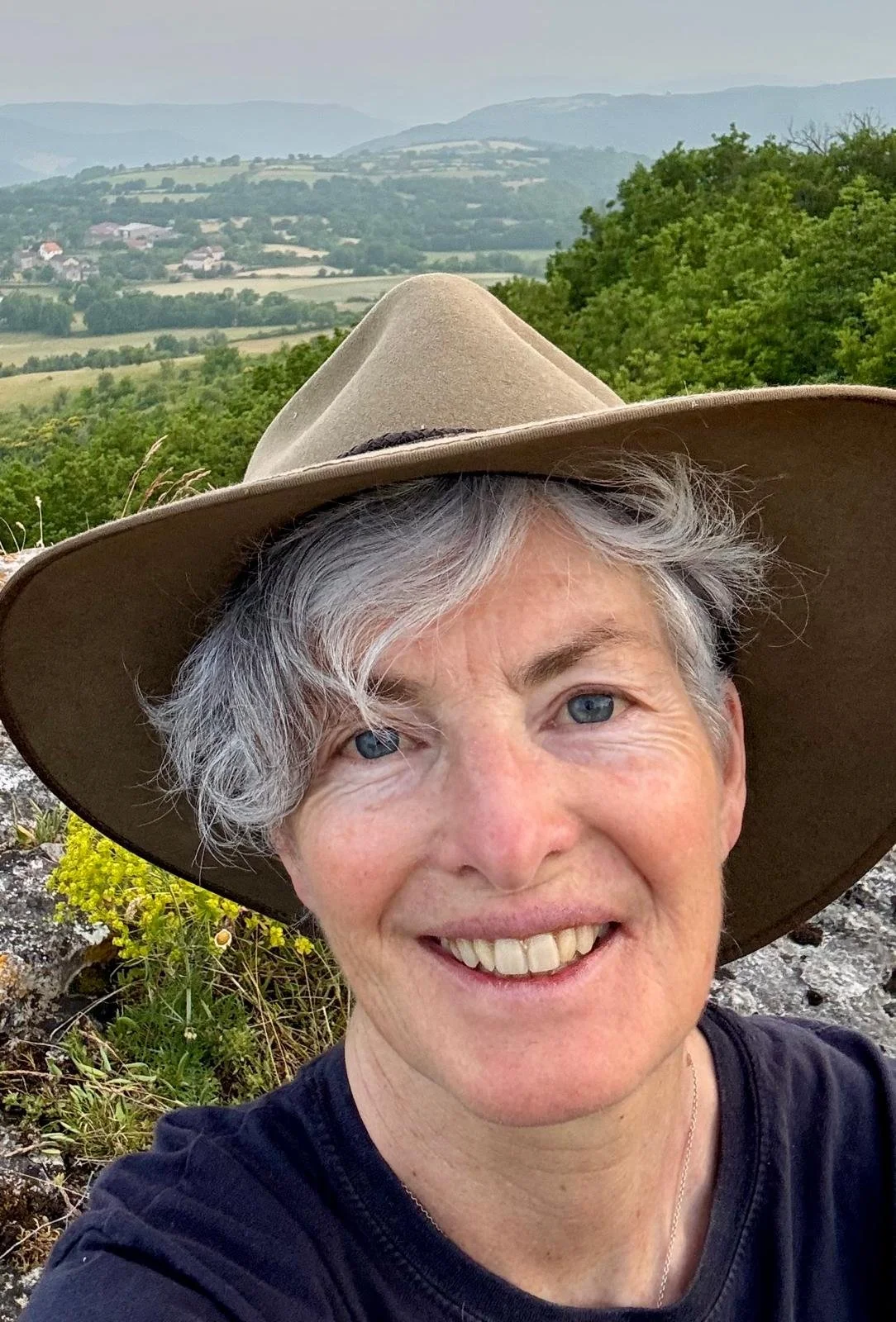 A smiling woman with short gray hair wearing a wide-brimmed beige hat taking a selfie outdoors with a landscape of rolling hills and trees in the background.