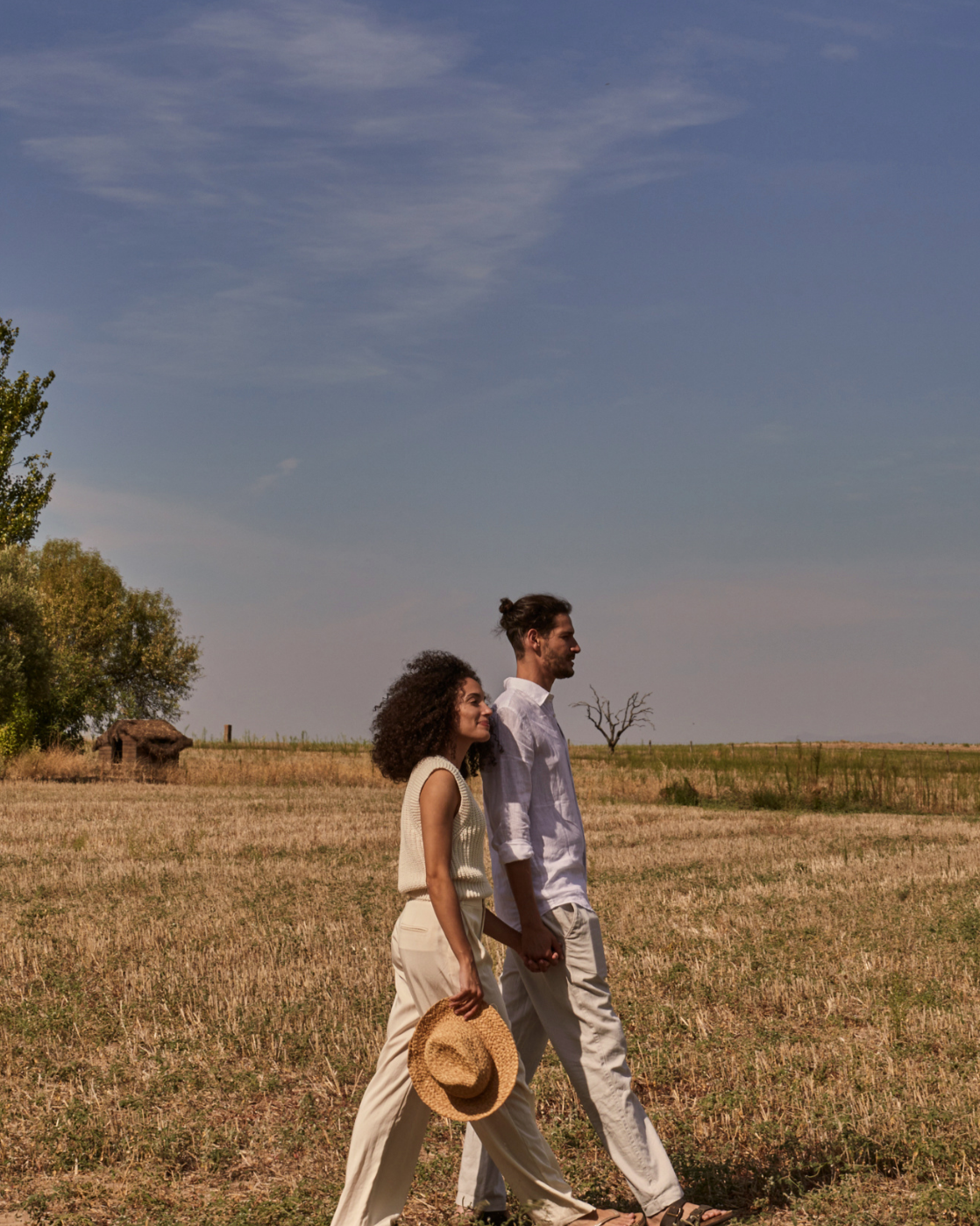 A happy couple walking hand in hand through a field under a blue sky, the woman holding a straw hat, both dressed in light-colored clothes.