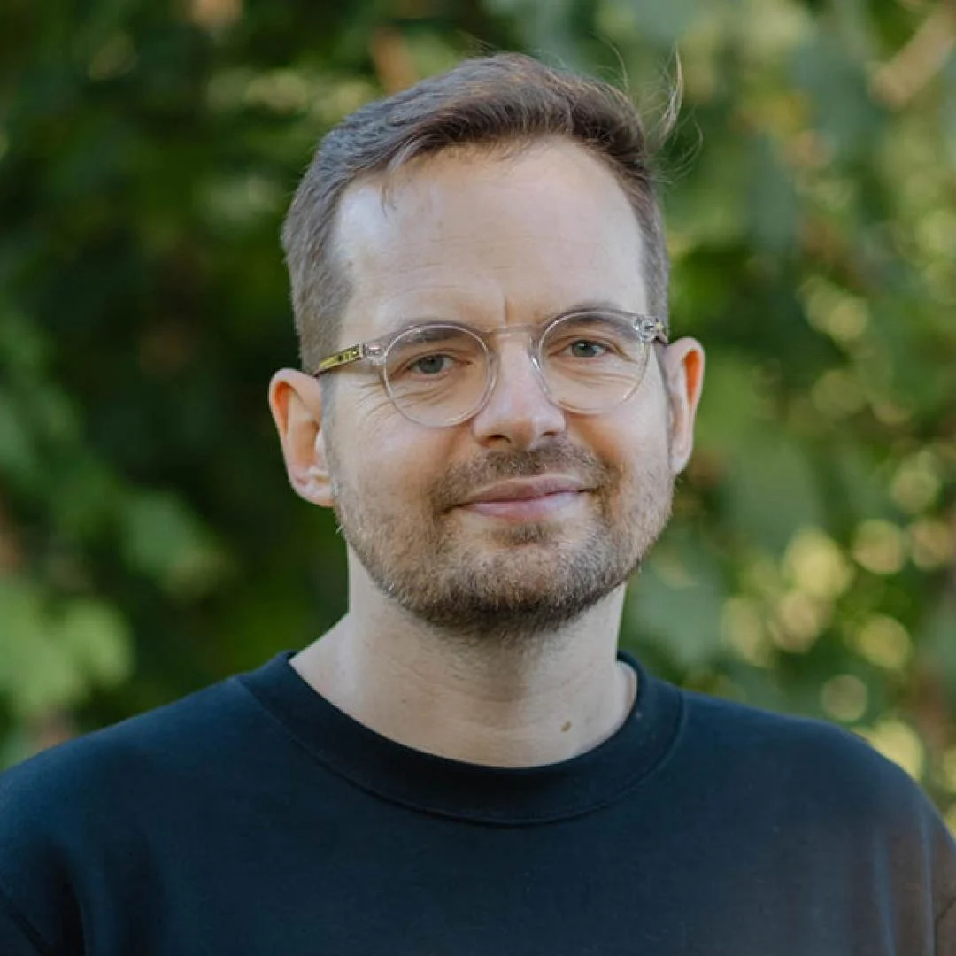 A man with glasses and a beard standing outdoors in front of green foliage.