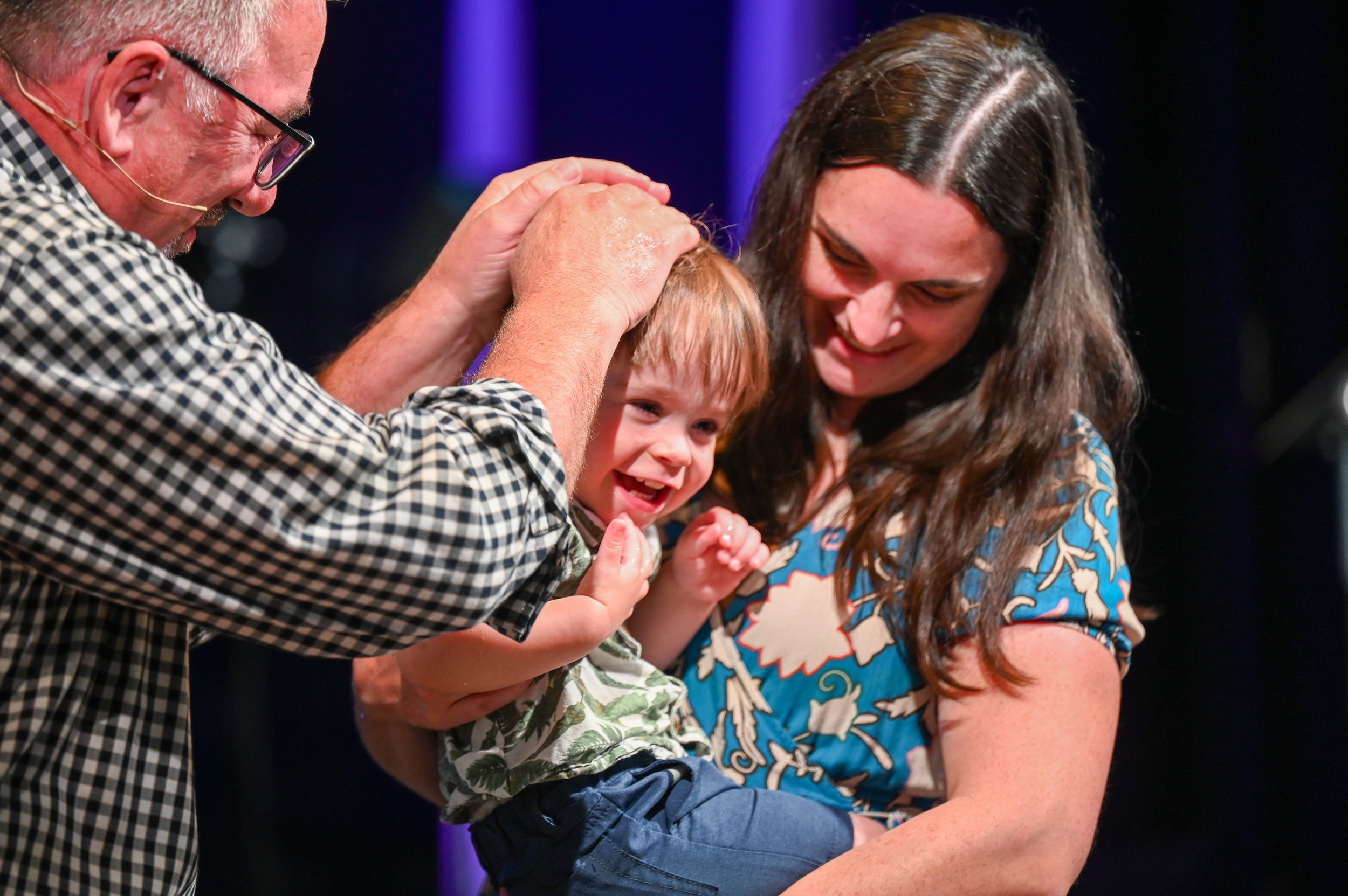 A joyful young boy being blessed or baptized by an older man, with a woman holding him, both smiling on a dark stage.