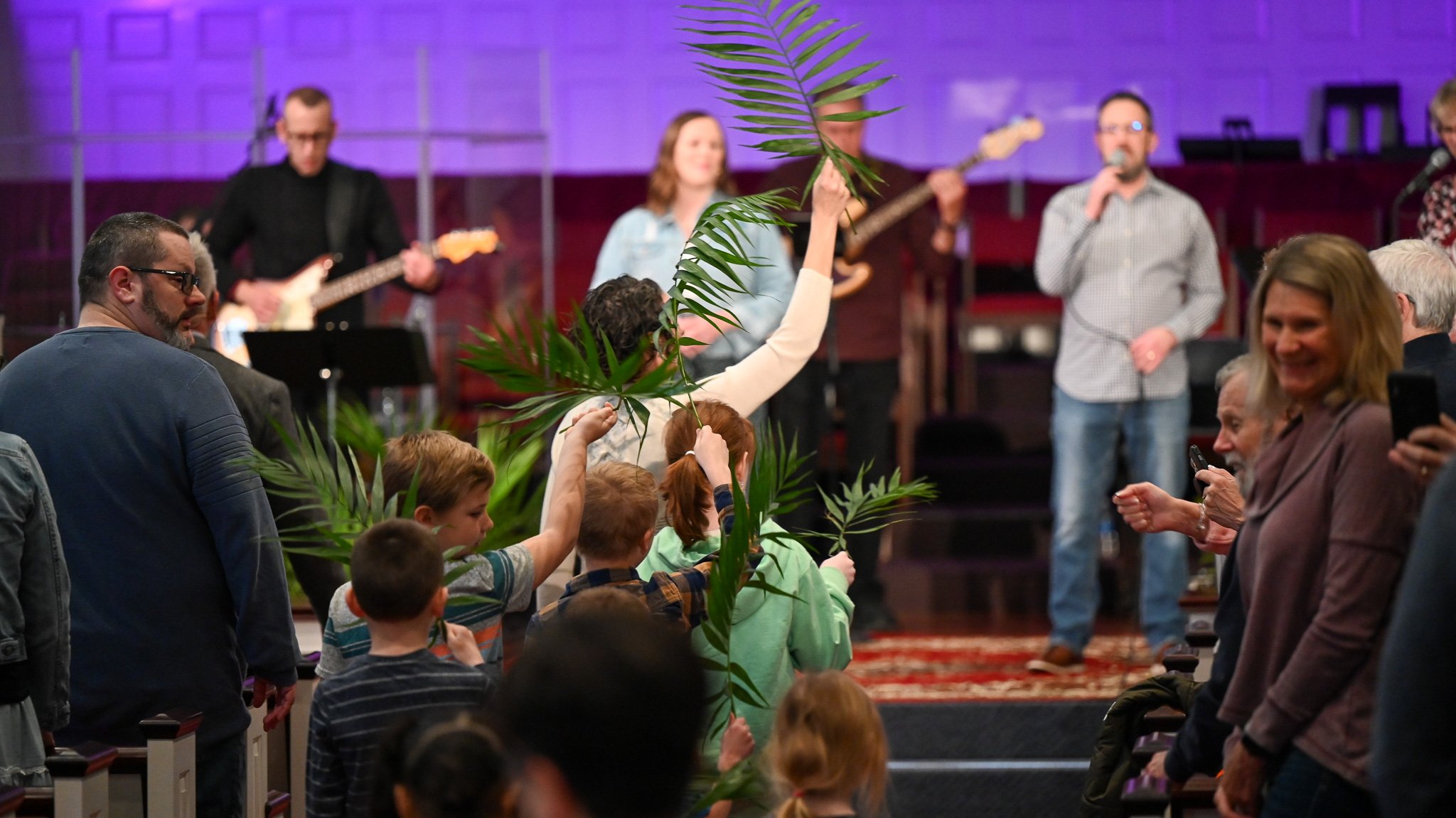 People attending a church service or event, with some holding large green palm leaves, while a band performs on stage in the background, illuminated by purple lighting.