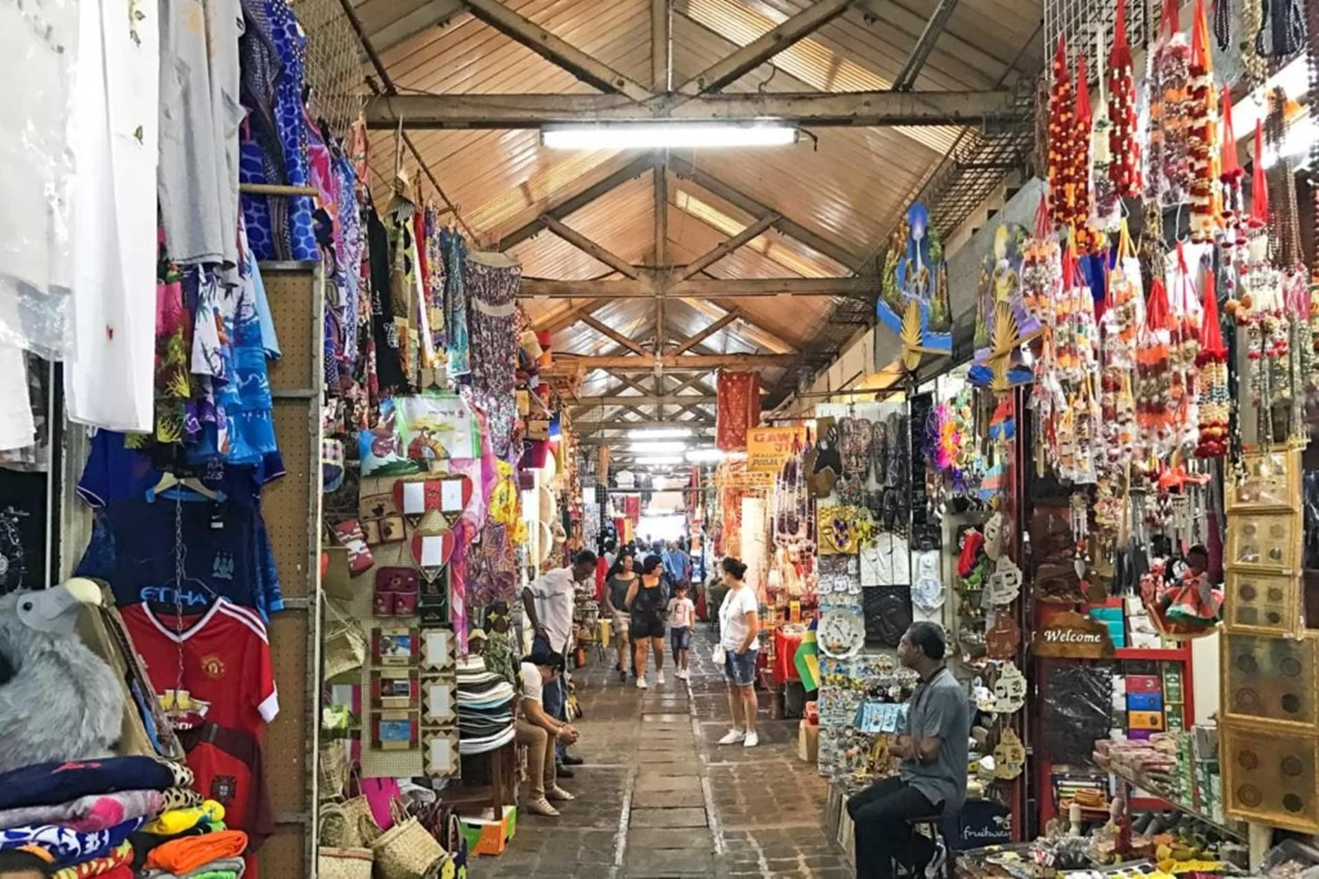 A bustling market with clothing, accessories, and souvenirs displayed on both sides of an aisle, with people shopping and browsing under a wooden roof with fluorescent lighting.